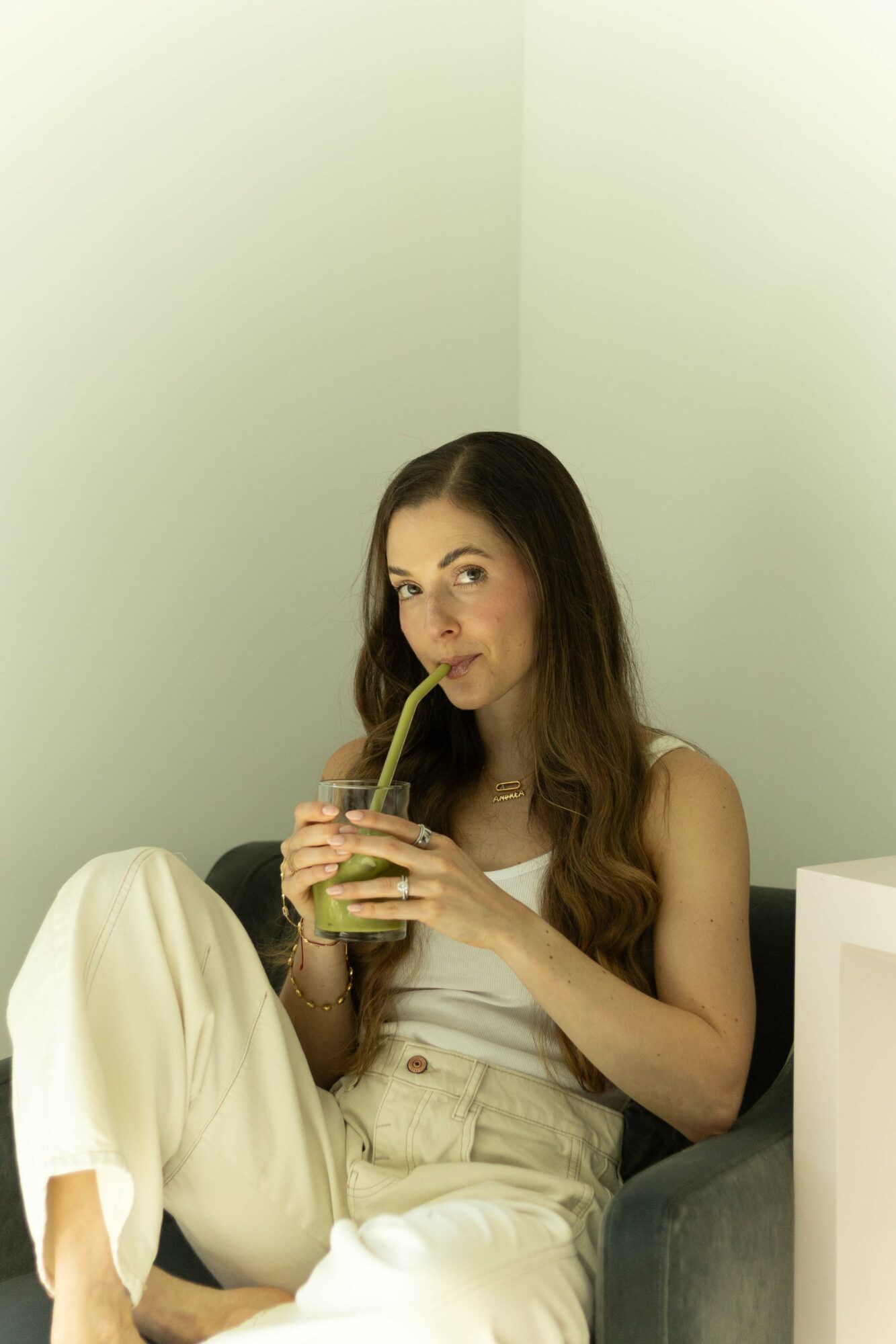 Young woman with long brown hair drinks from a glass with a straw, sitting on a couch in a light-colored room.