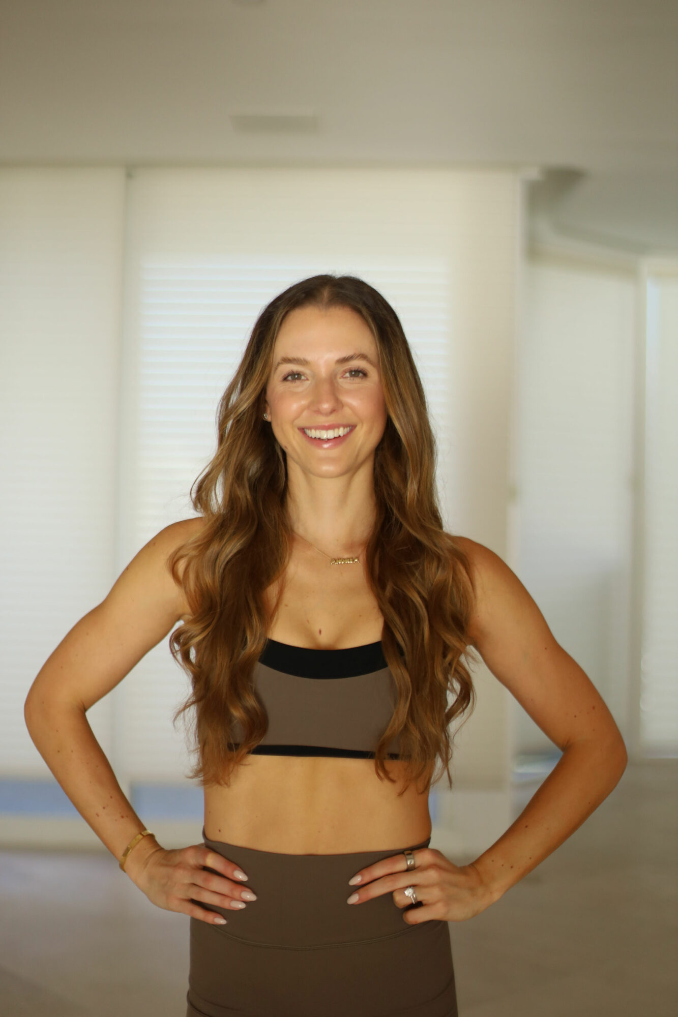Smiling woman with long wavy hair in a sports bra and leggings, hands on hips, indoors with windows behind.