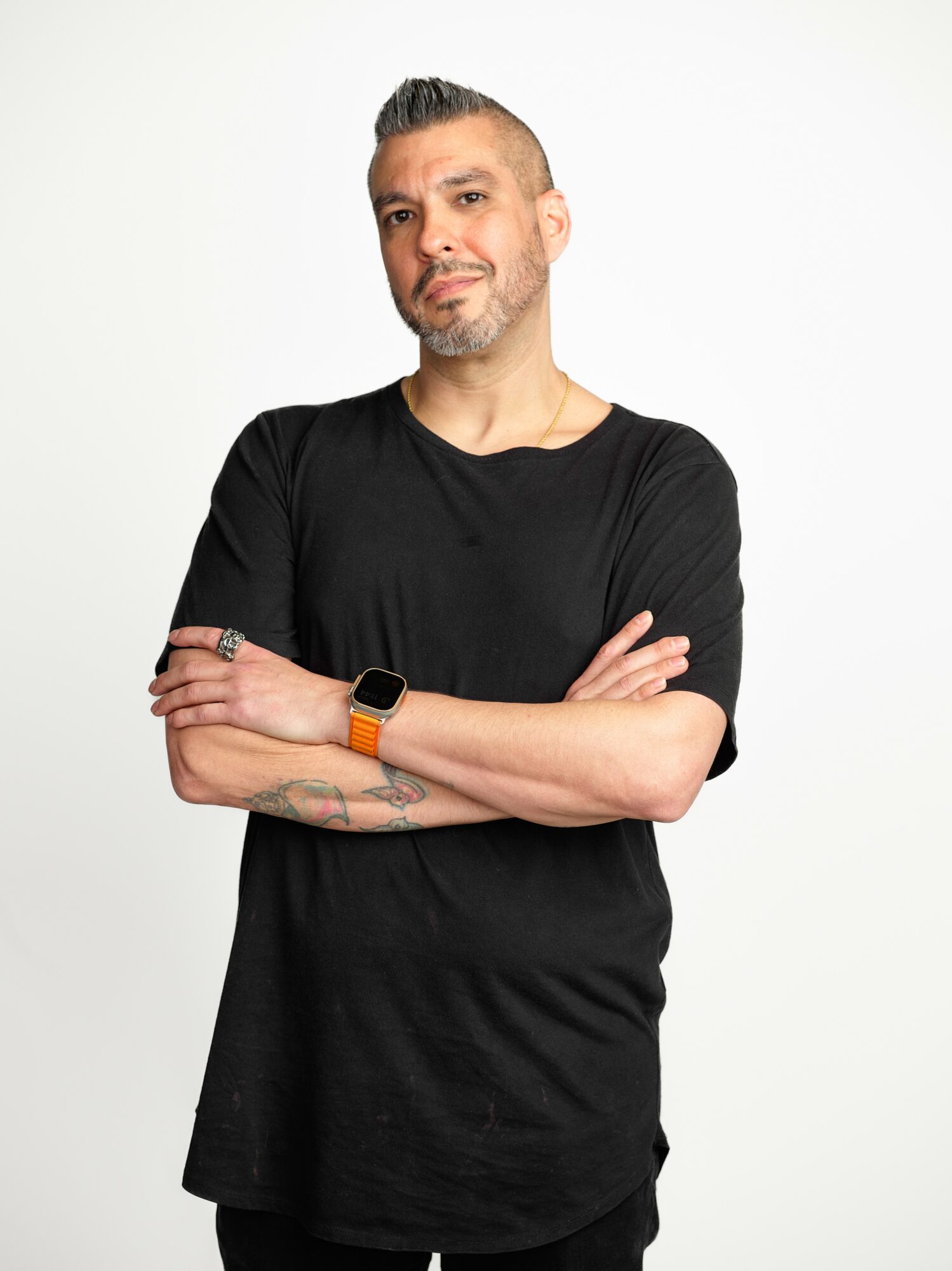 Man with short hair and beard, wearing a black t-shirt and smartwatch, crossing arms, standing against a white background.