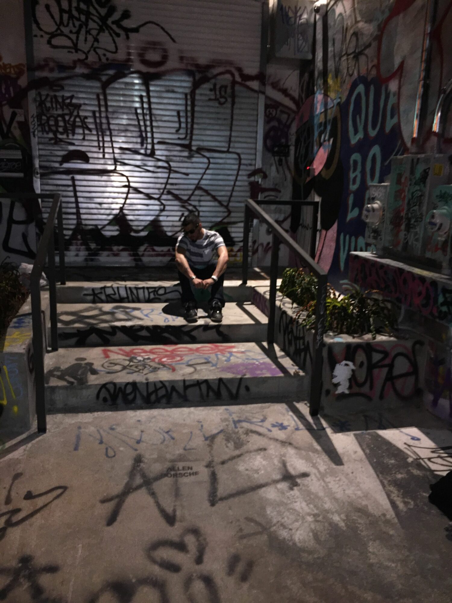 Person sitting on a bench in front of graffiti-covered walls and railings, with shadows on the ground.