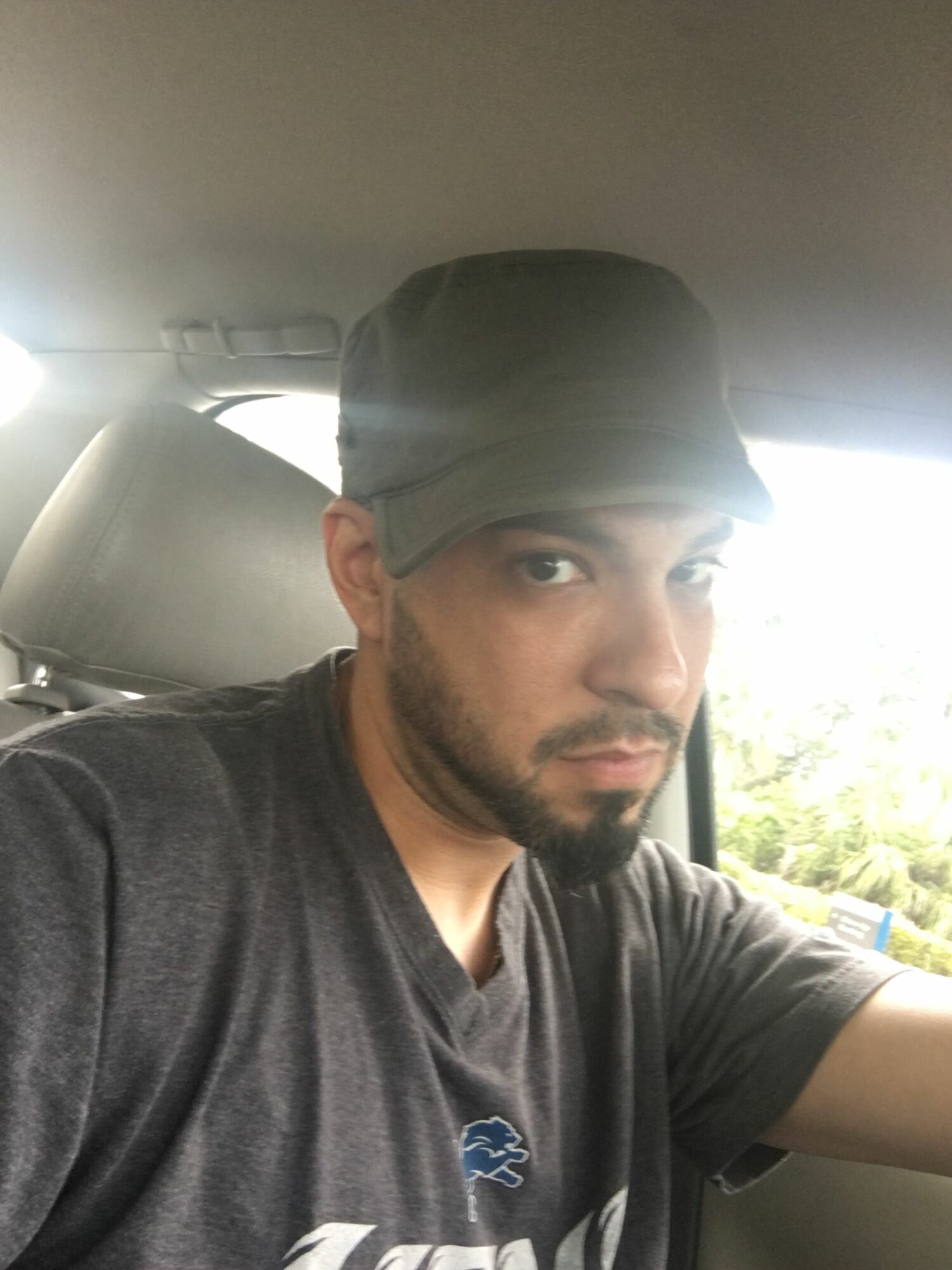 Man with a beard and cap sitting in a car, looking at the camera, with sunlight outside the window.