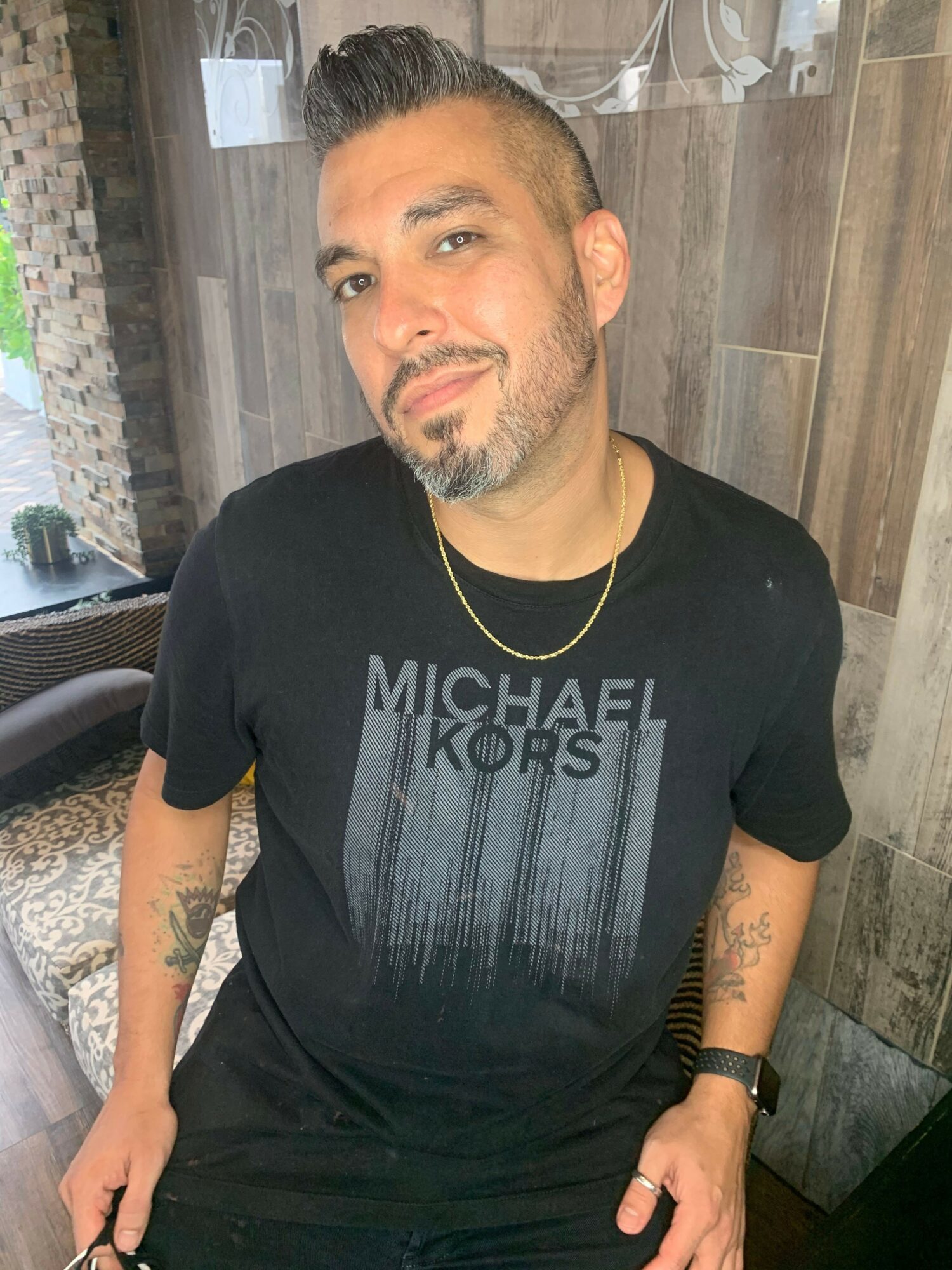 Man with dark hair, beard, and tattoos wearing a black Michael Kors T-shirt, standing indoors against a wooden wall.