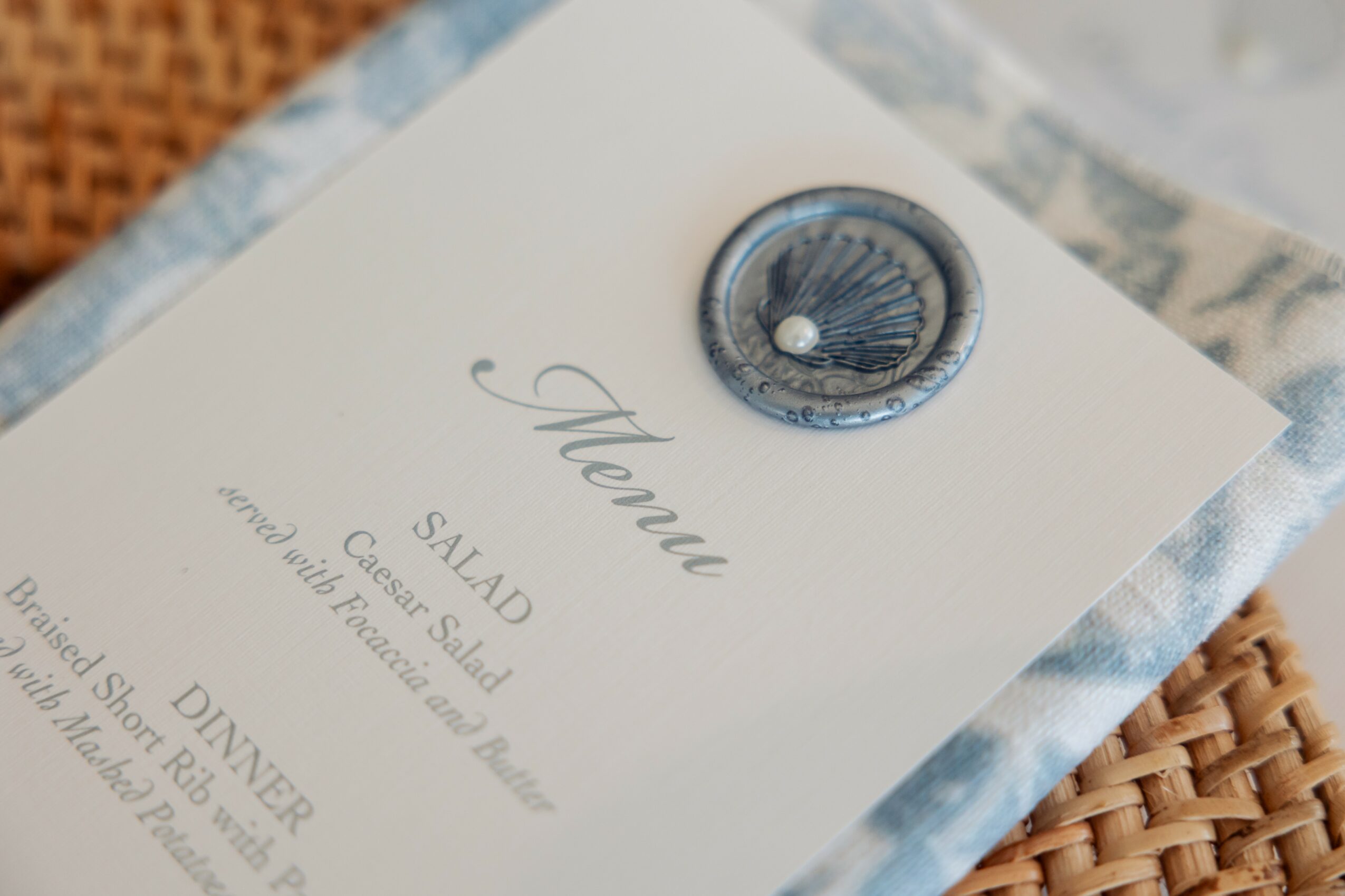 Close-up of a menu with a decorative button on top, placed on a woven surface, with a blurred background.