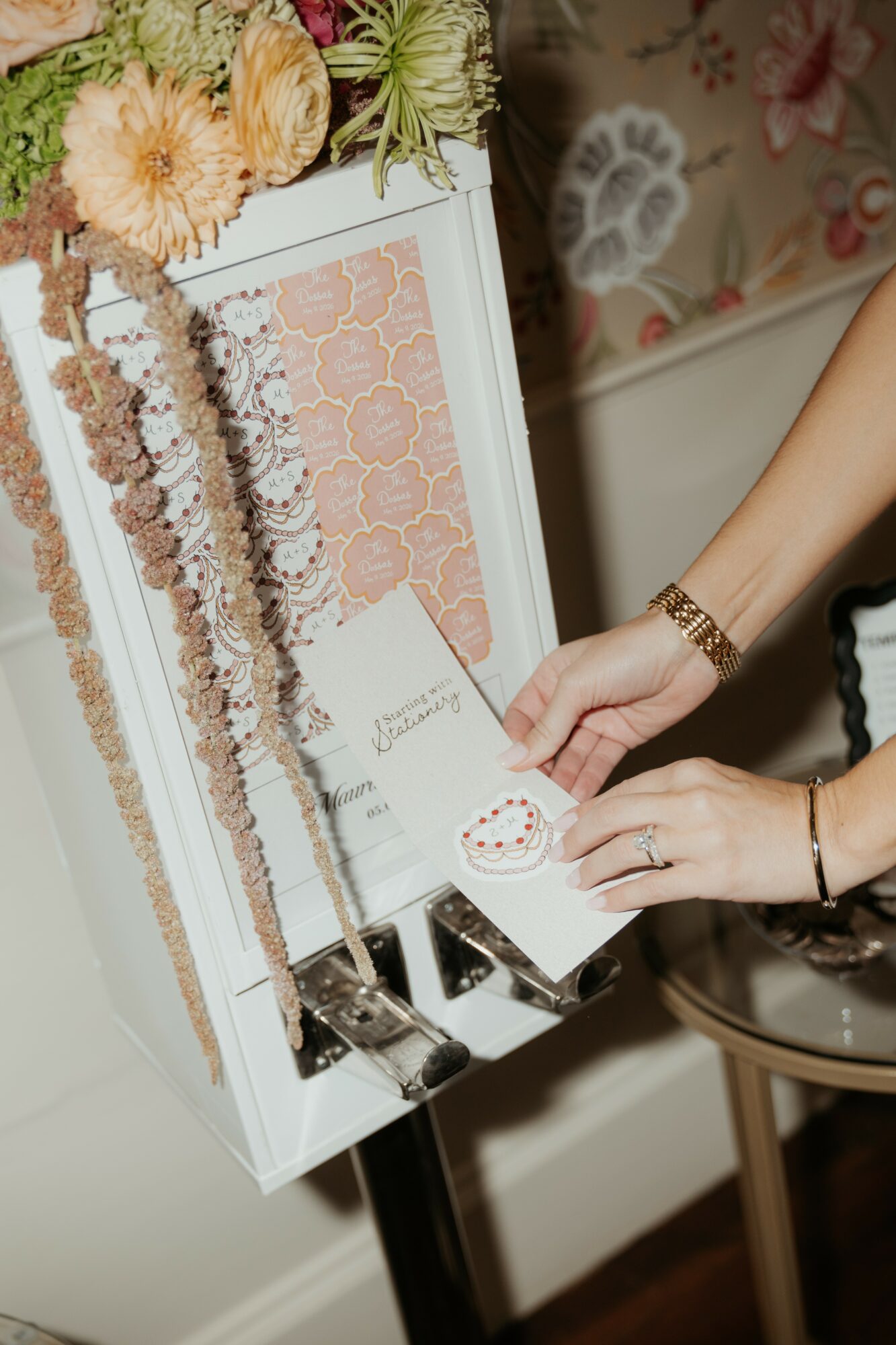 Person's hand with a bracelet reaches for a card in a decorated display box with flowers and patterned paper, indoors.