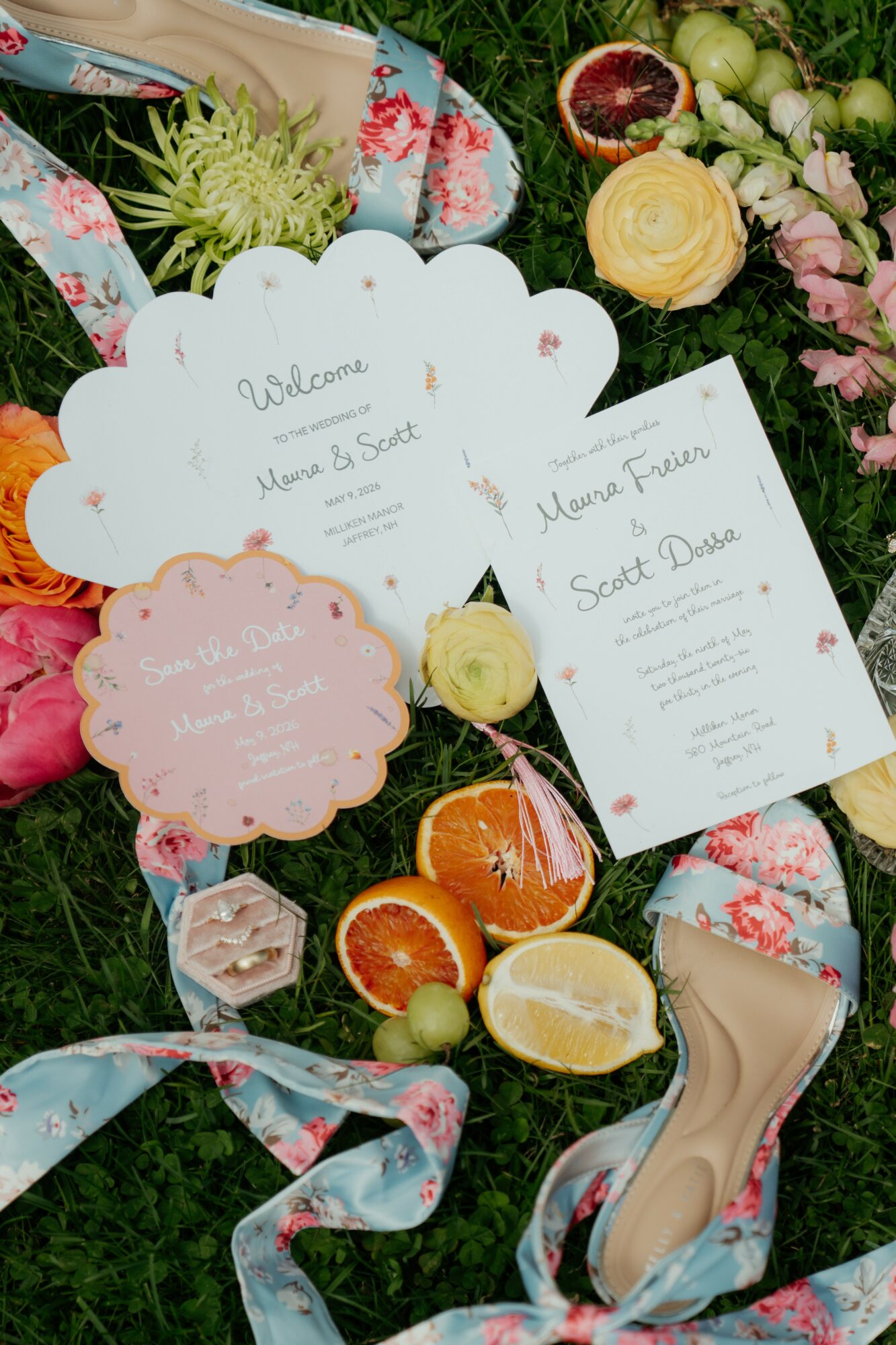 Wedding invitation cards, flowers, and a high heel shoe arranged on grass, with citrus fruits and decorative elements.