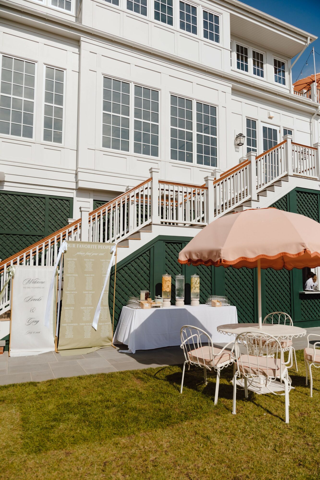 Outdoor patio with tables, chairs, and a large pink umbrella in front of a white building with large windows.
