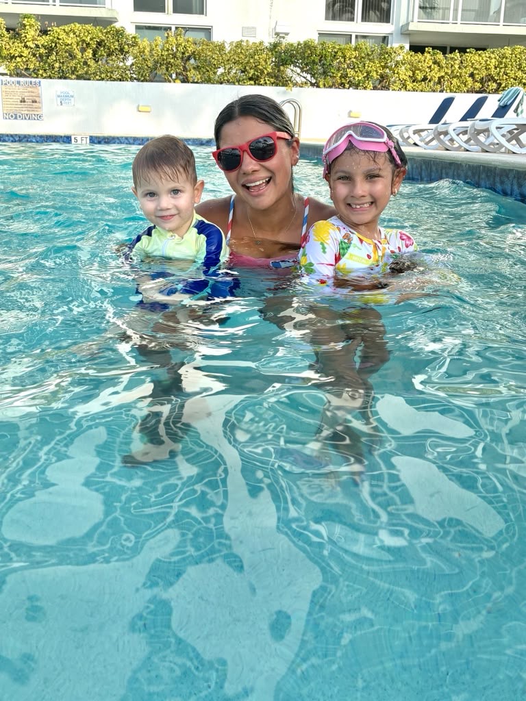 Woman with two children in a swimming pool, all smiling, wearing goggles and swimwear, sunny day, poolside chairs in background.
