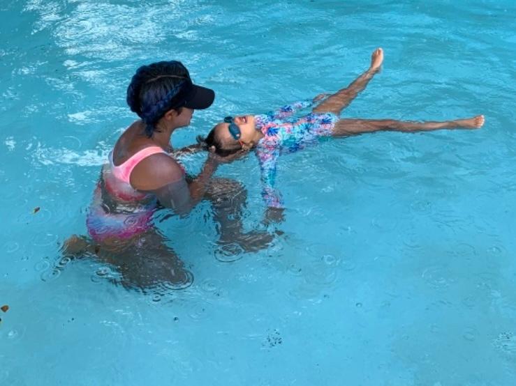 Two people in a swimming pool, one holding the other, who is floating on their back with arms and legs extended.