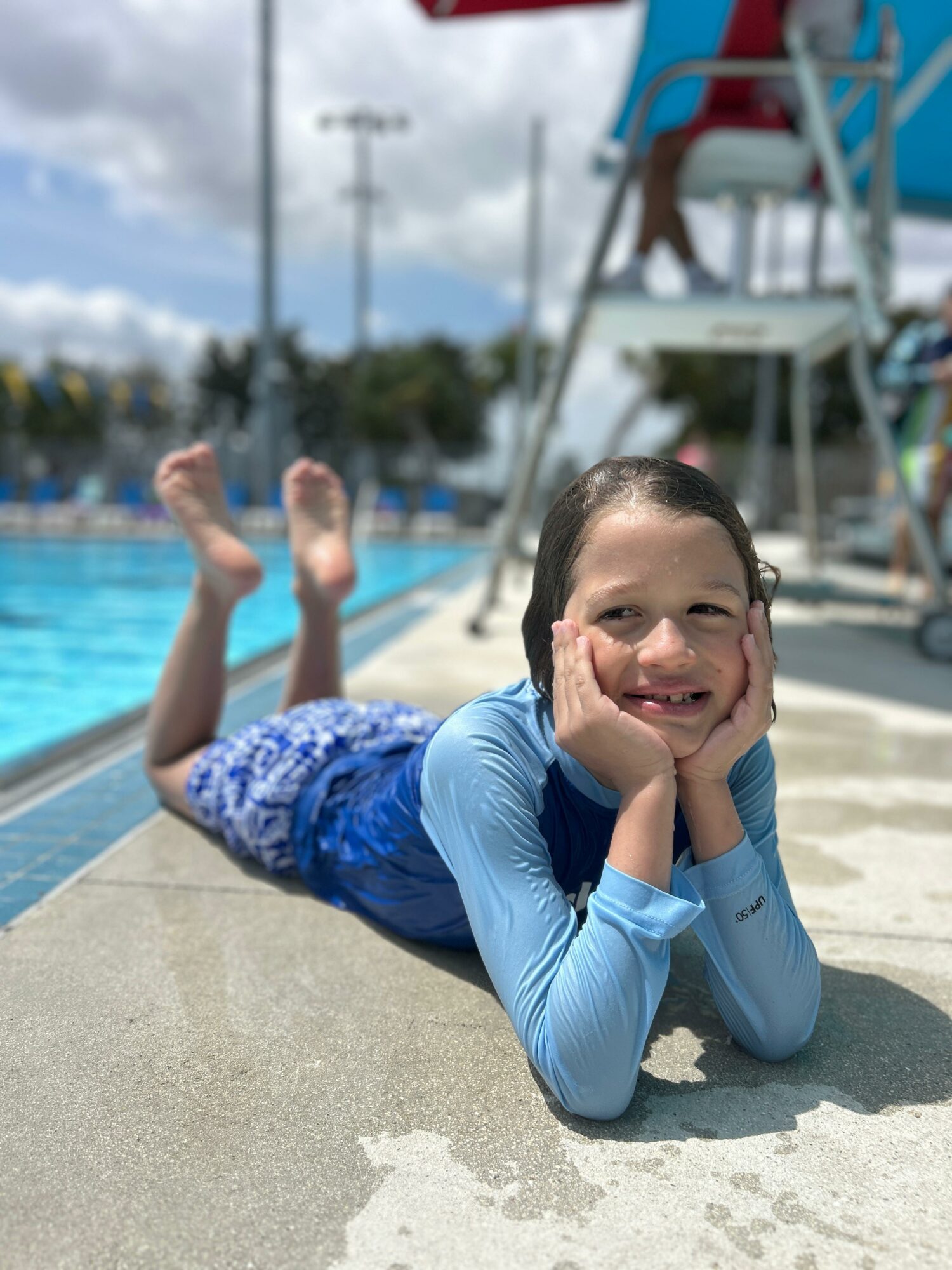 Child lying on poolside with feet in the air, smiling, wearing a blue swimsuit, near a lifeguard chair and pool.