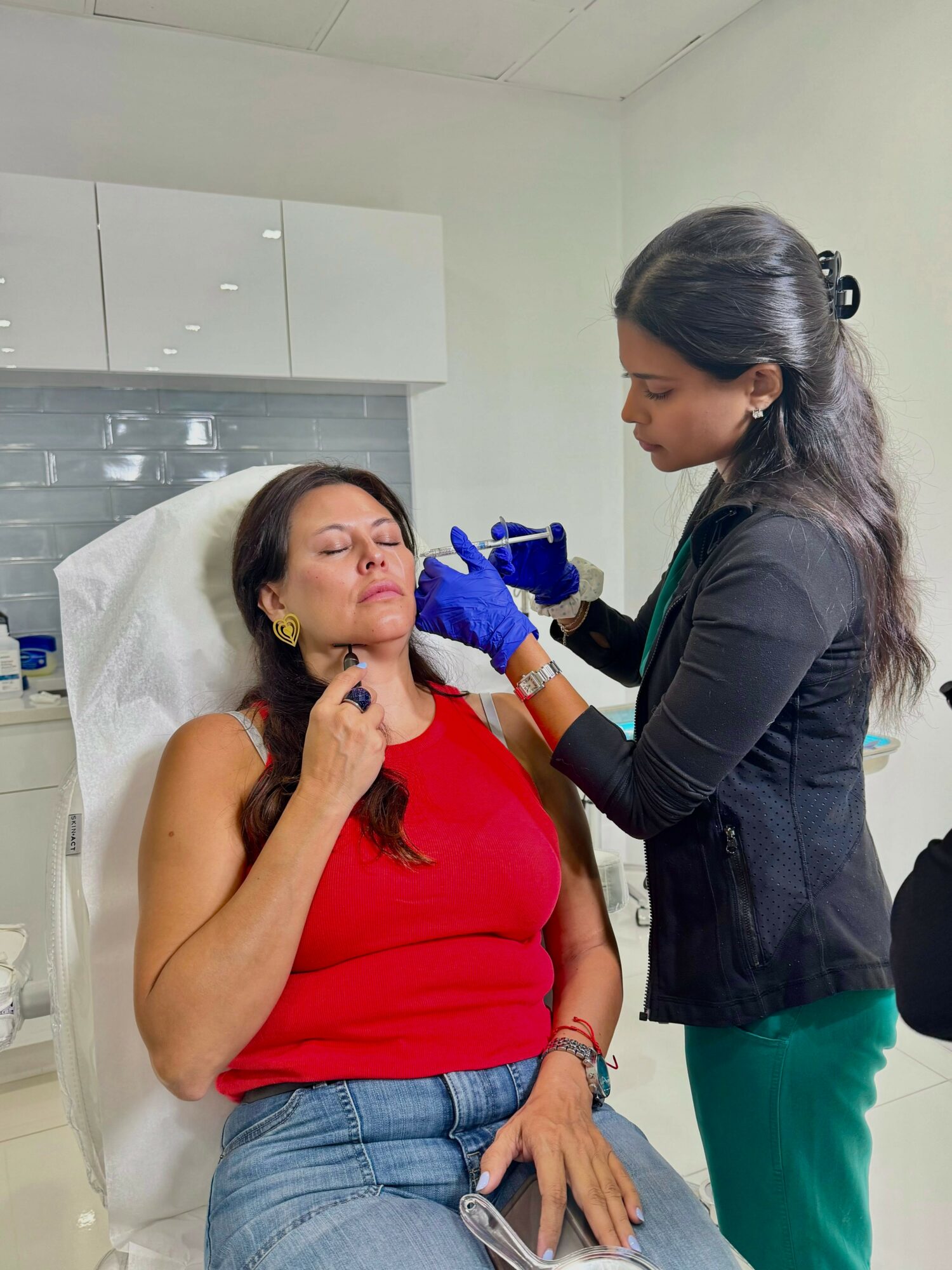 A woman in a red top is receiving a cosmetic treatment from a professional in a clinical setting.