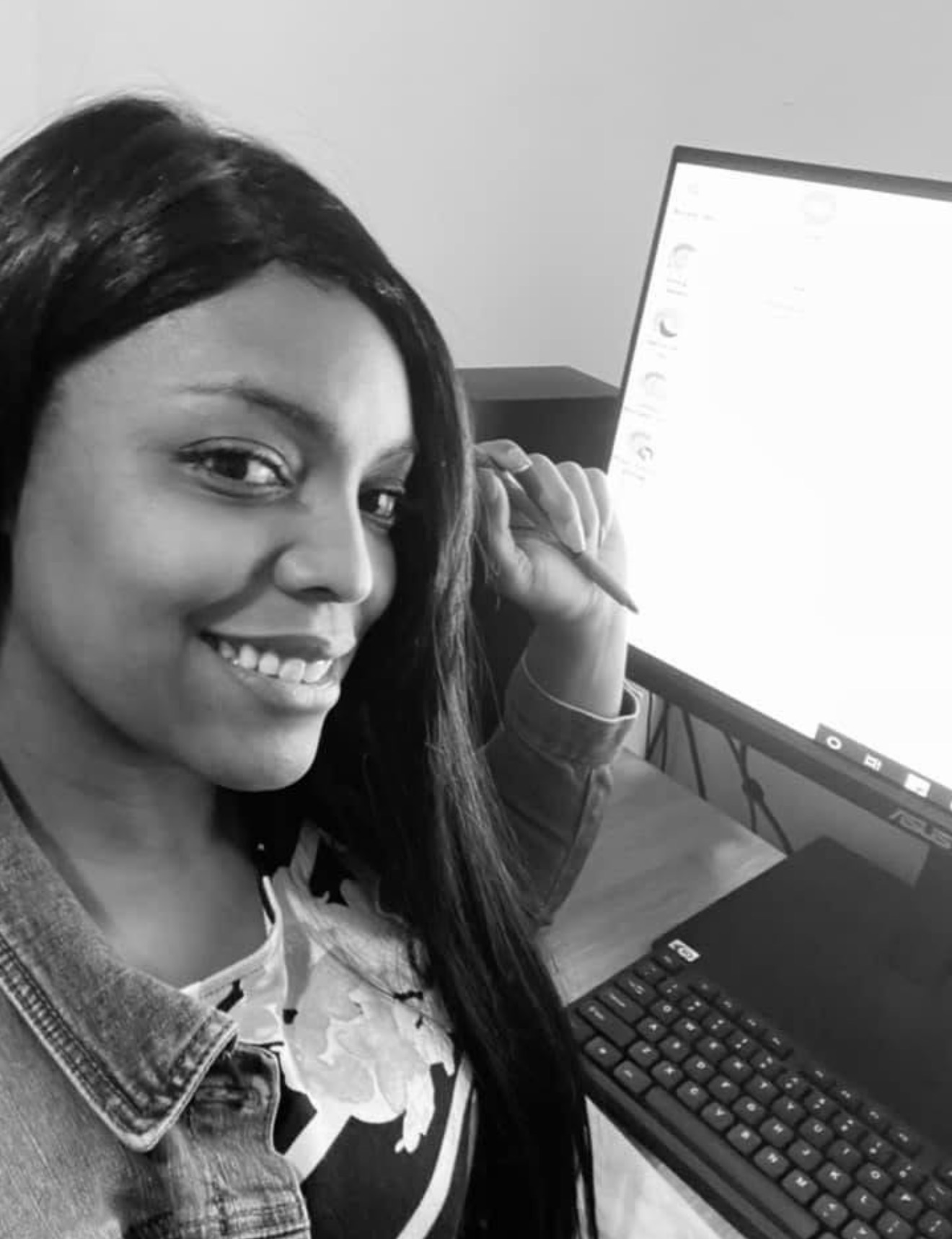 Young woman with long hair smiling at computer monitor in grayscale, sitting at desk with keyboard and mouse.