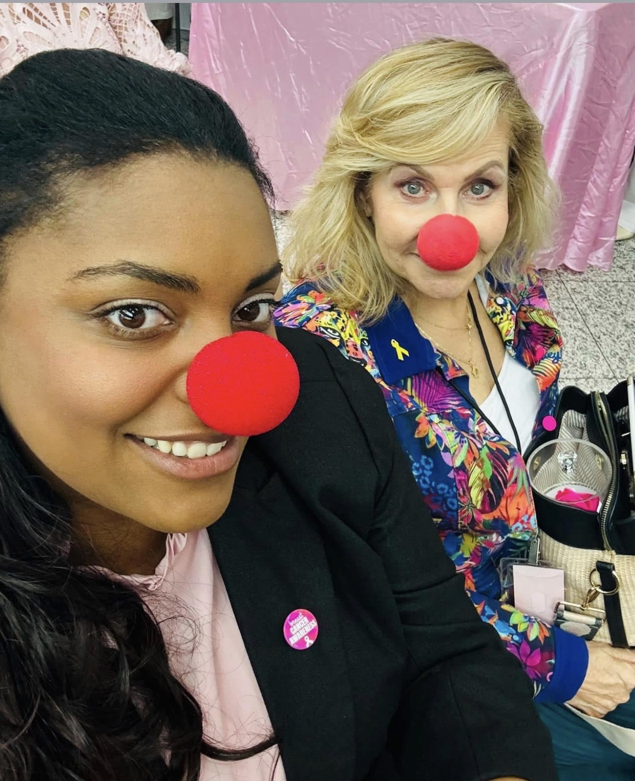 Two women wearing red clown noses, smiling, sitting indoors with pink and gray backgrounds.