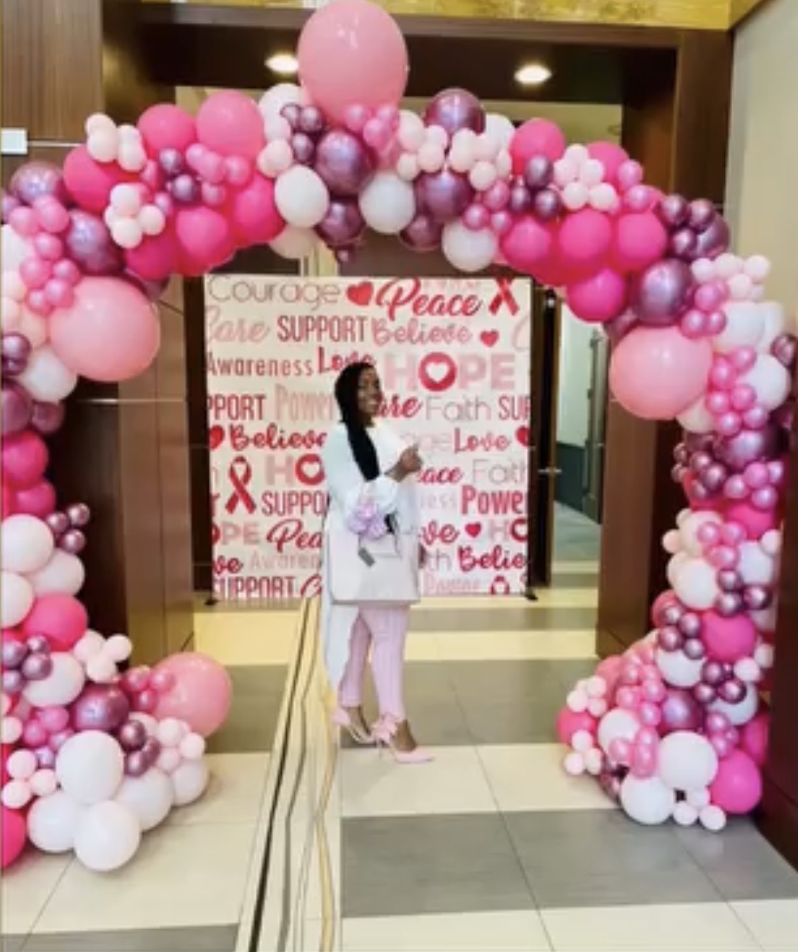 Person standing under a pink and white balloon arch with a backdrop of words like love, hope, and support, in an indoor setting.