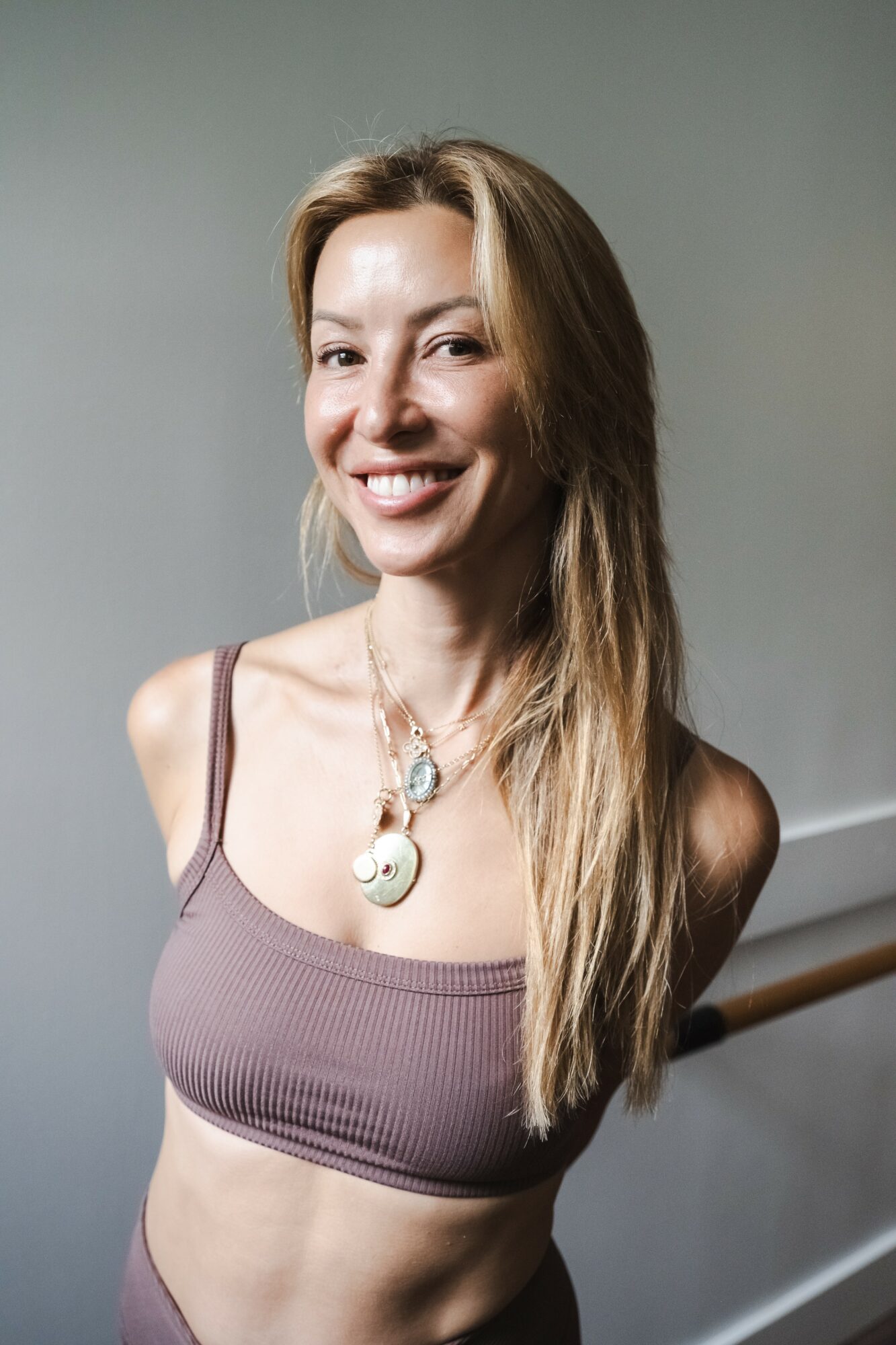 Smiling woman with long hair wearing a sleeveless top and necklace, standing indoors against a plain background.
