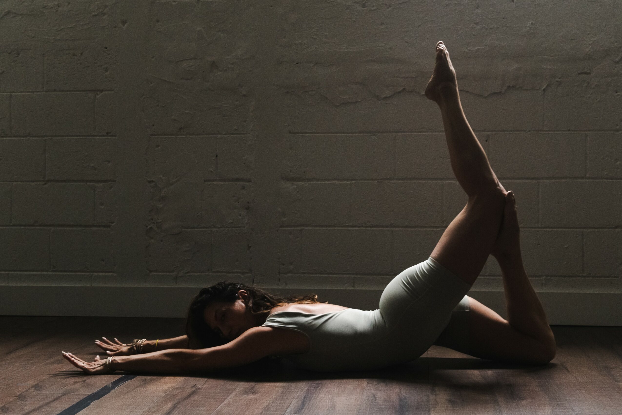 Person performing a yoga pose on the floor with a brick wall background, stretching forward with one leg bent and the other extended upward.