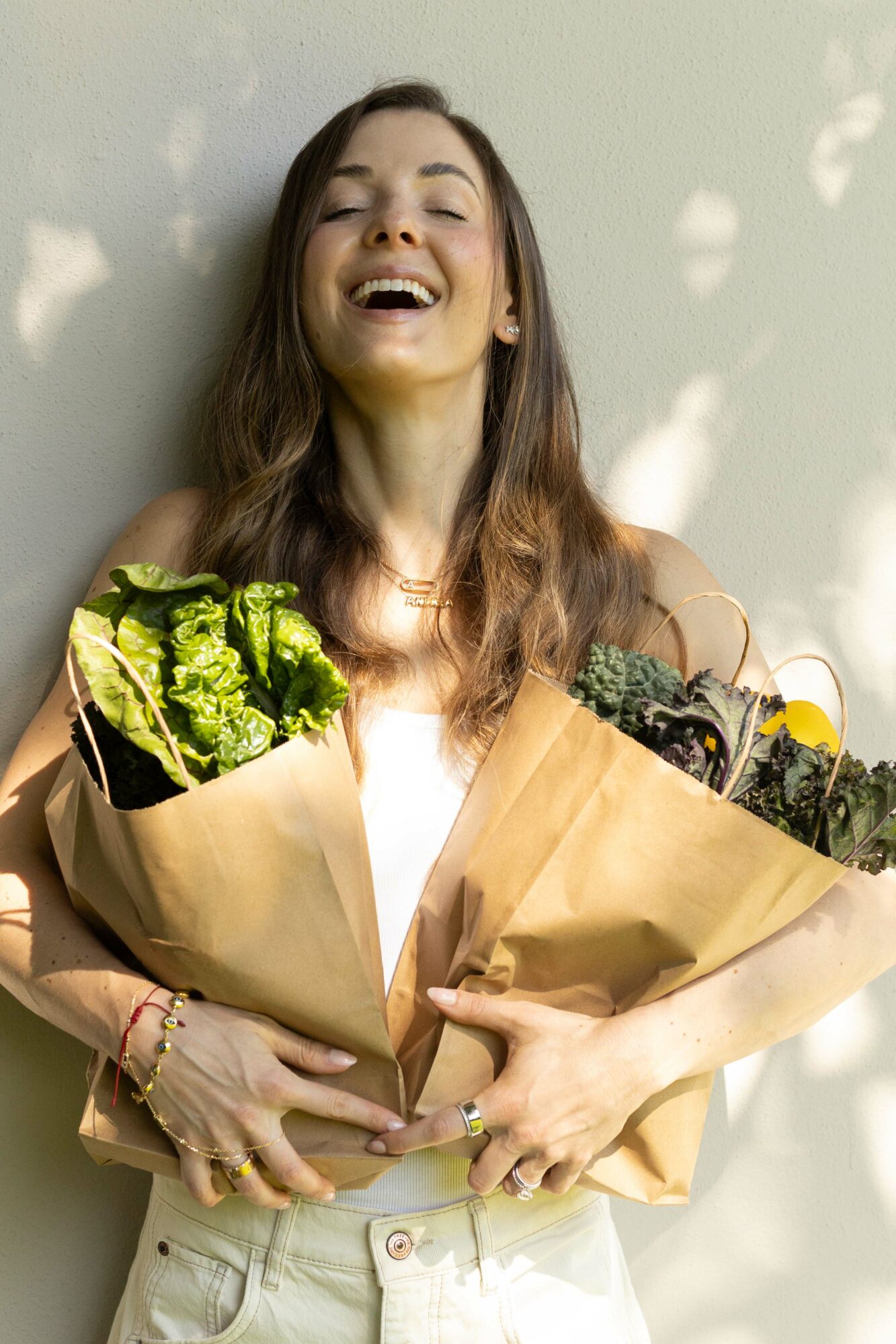 Smiling woman holding two bouquets of flowers, standing against a light-colored wall with shadows, wearing a white top and light pants.