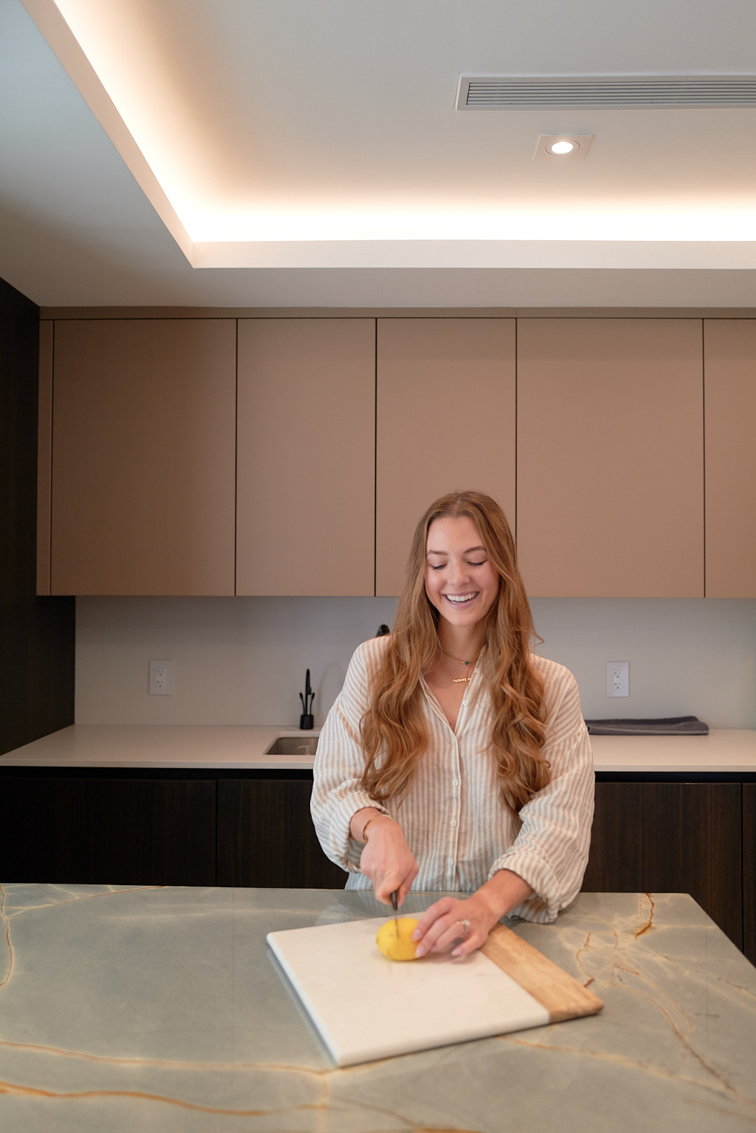 Young woman with long hair smiling while cutting a lemon on a cutting board in a modern kitchen.