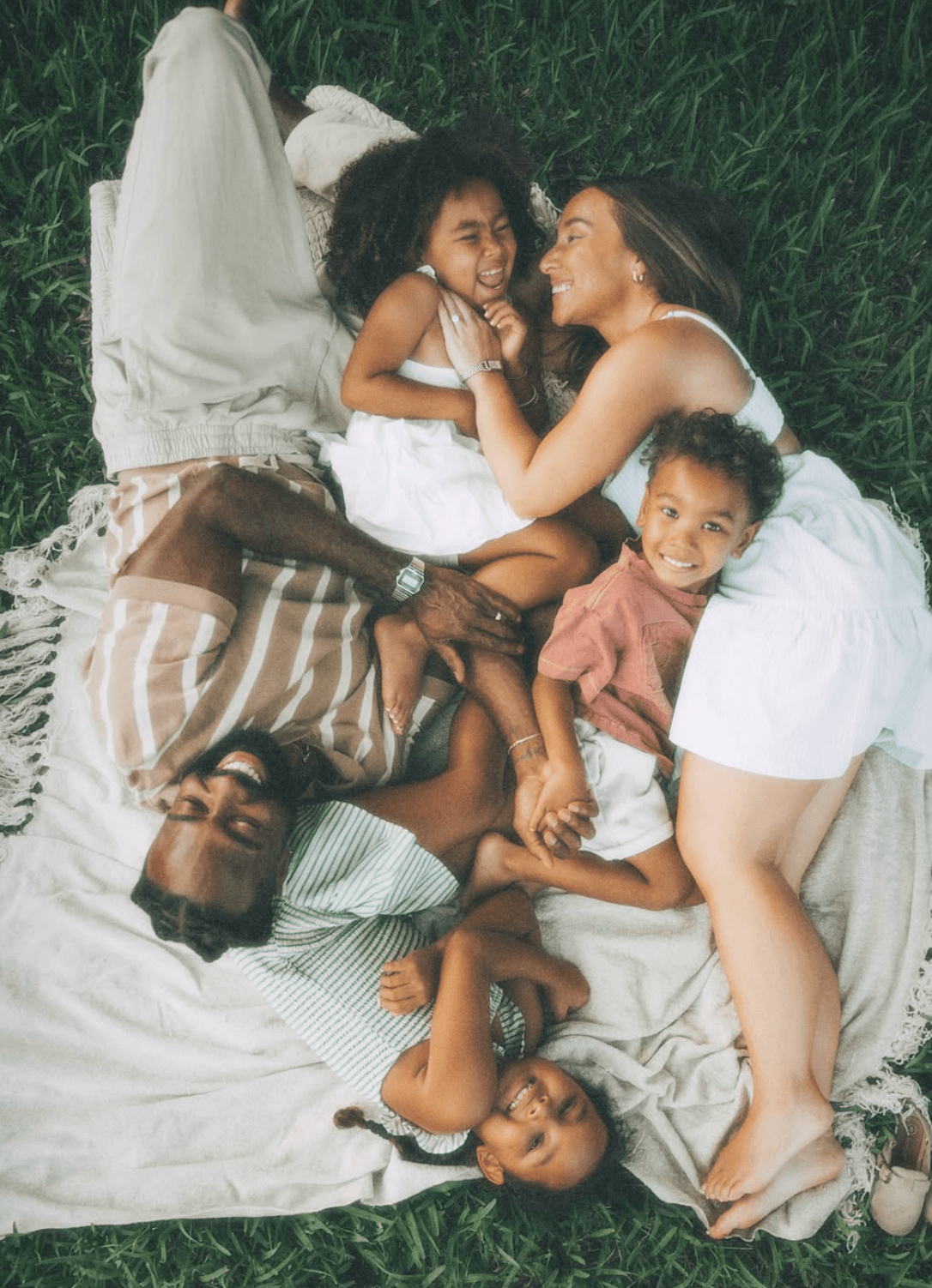 Group of five people lying on grass, smiling and interacting, with two children and three adults, outdoors.