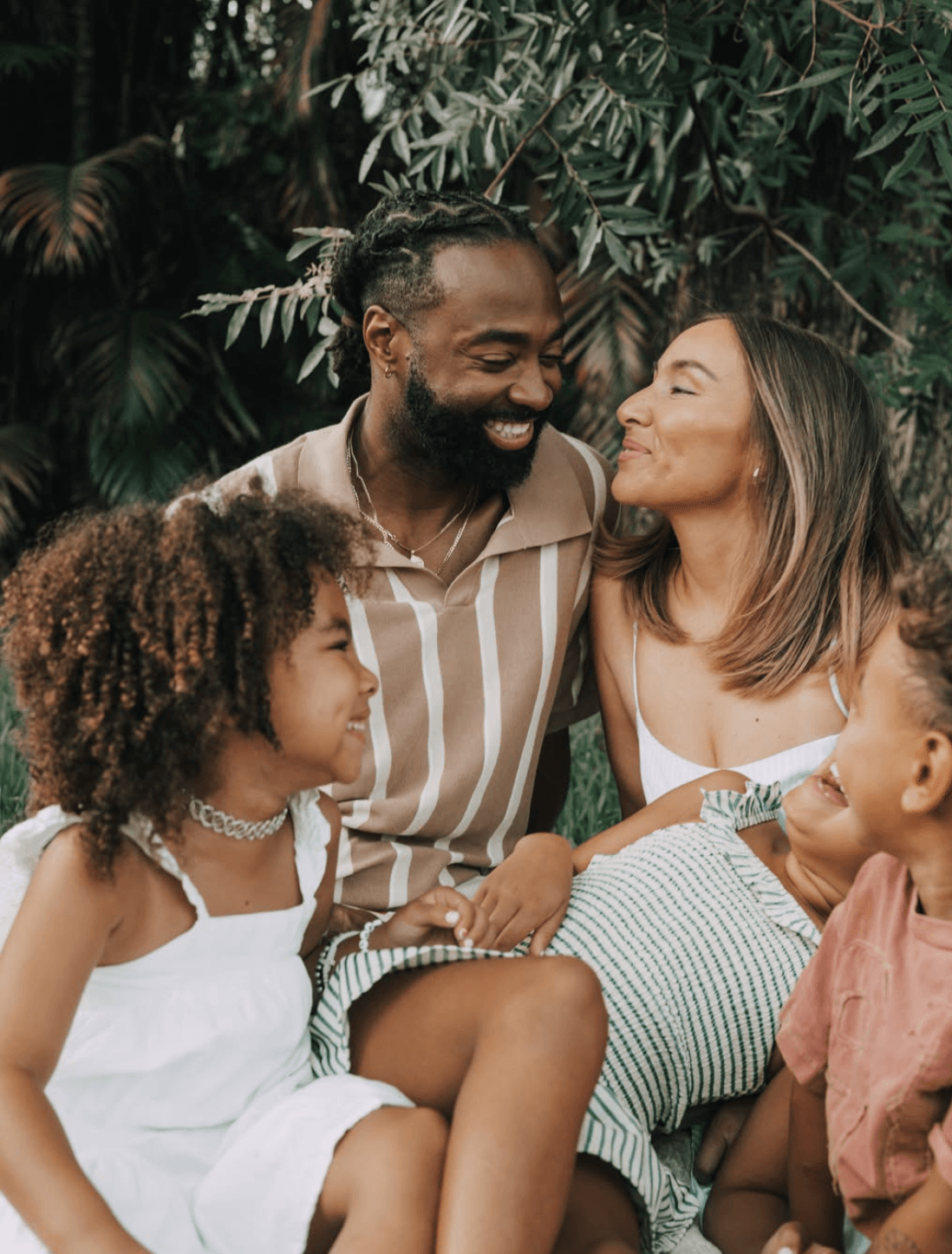 Four people, two adults and two children, smiling and interacting outdoors among green foliage.