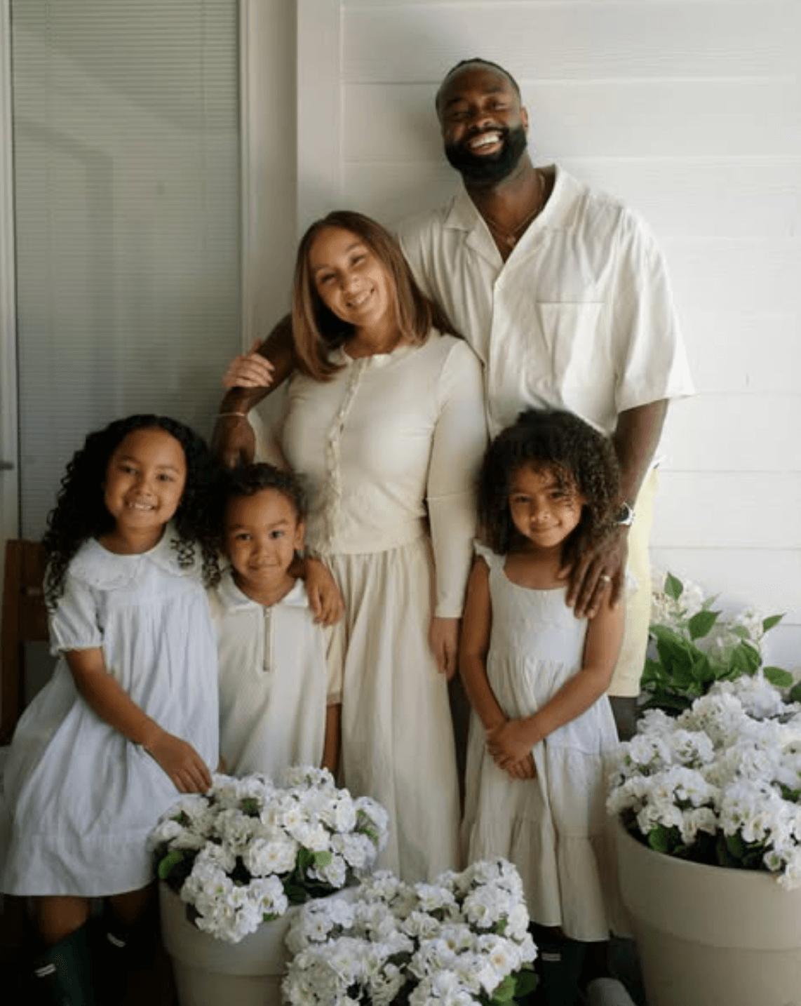 Group of five people, two adults and three children, standing indoors with white flowers in front, smiling, in light-colored clothing.