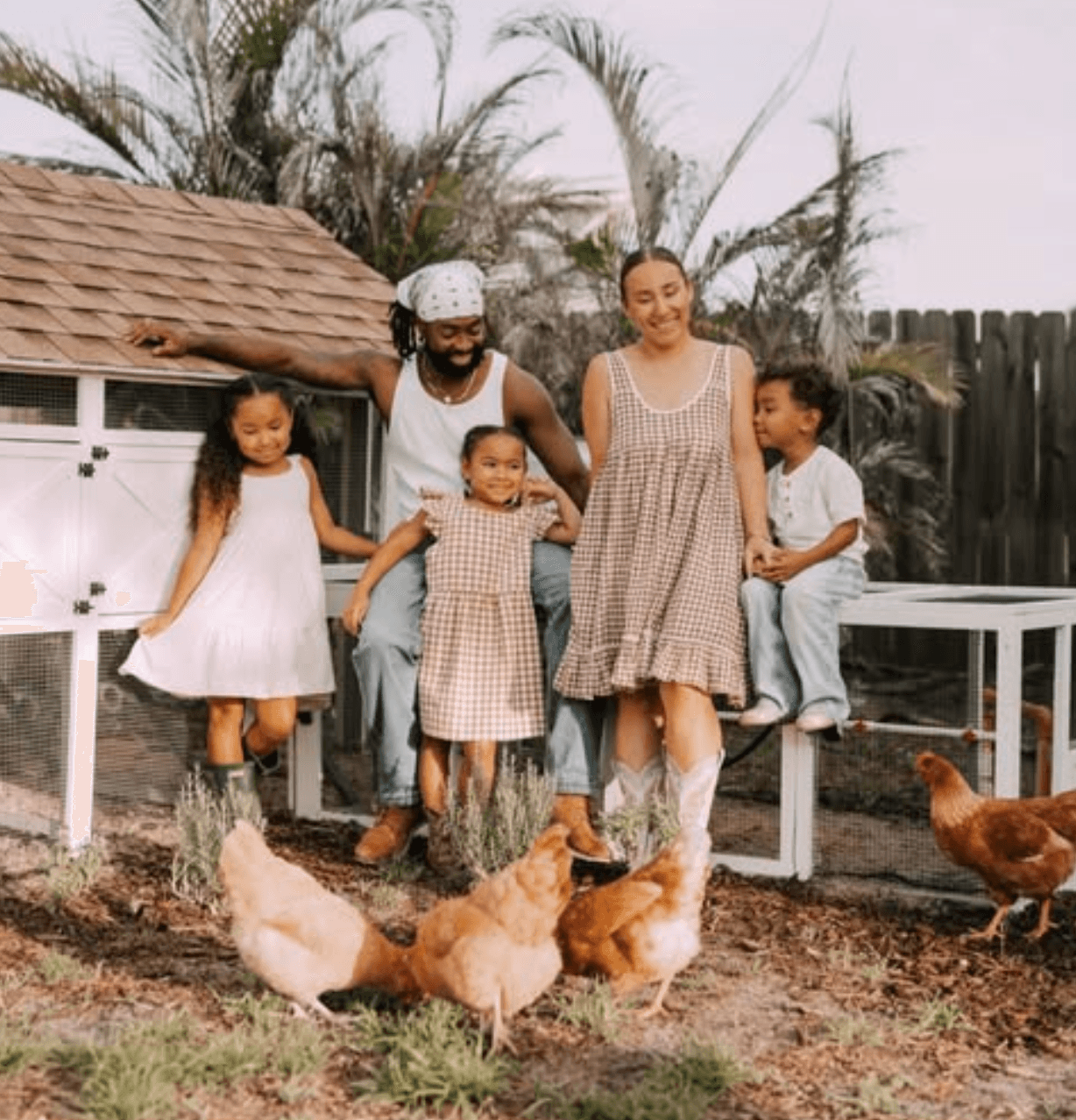 Family with children and chickens outdoors, smiling, standing on dirt ground, wooden fence and trees in background.