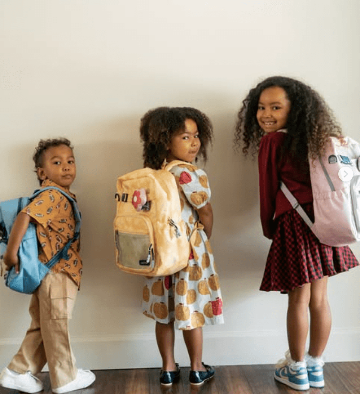 Three children with backpacks standing against a plain wall, smiling and looking at the camera.