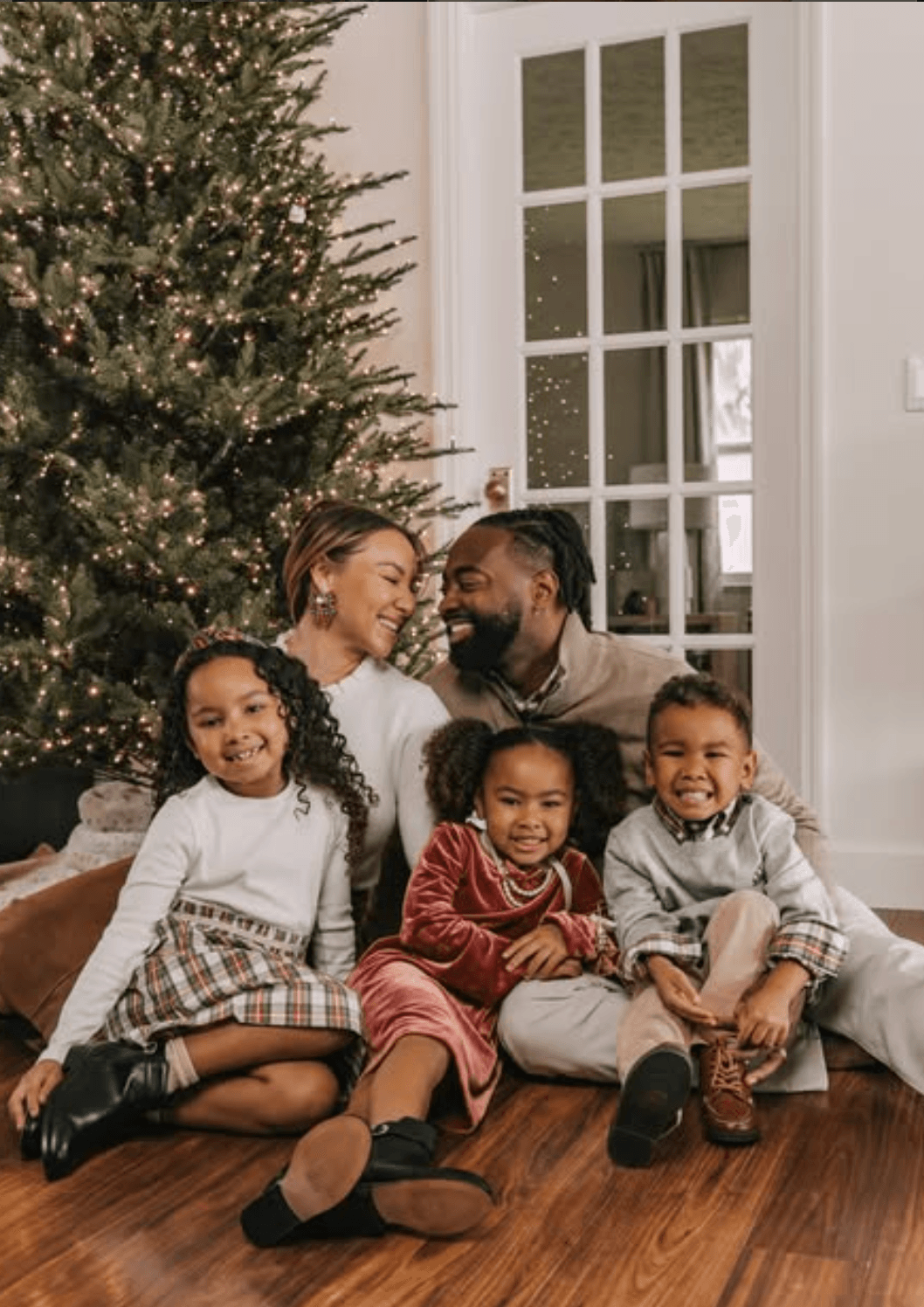 Family of five sitting on wooden floor near Christmas tree, smiling, indoors with large window and white walls.