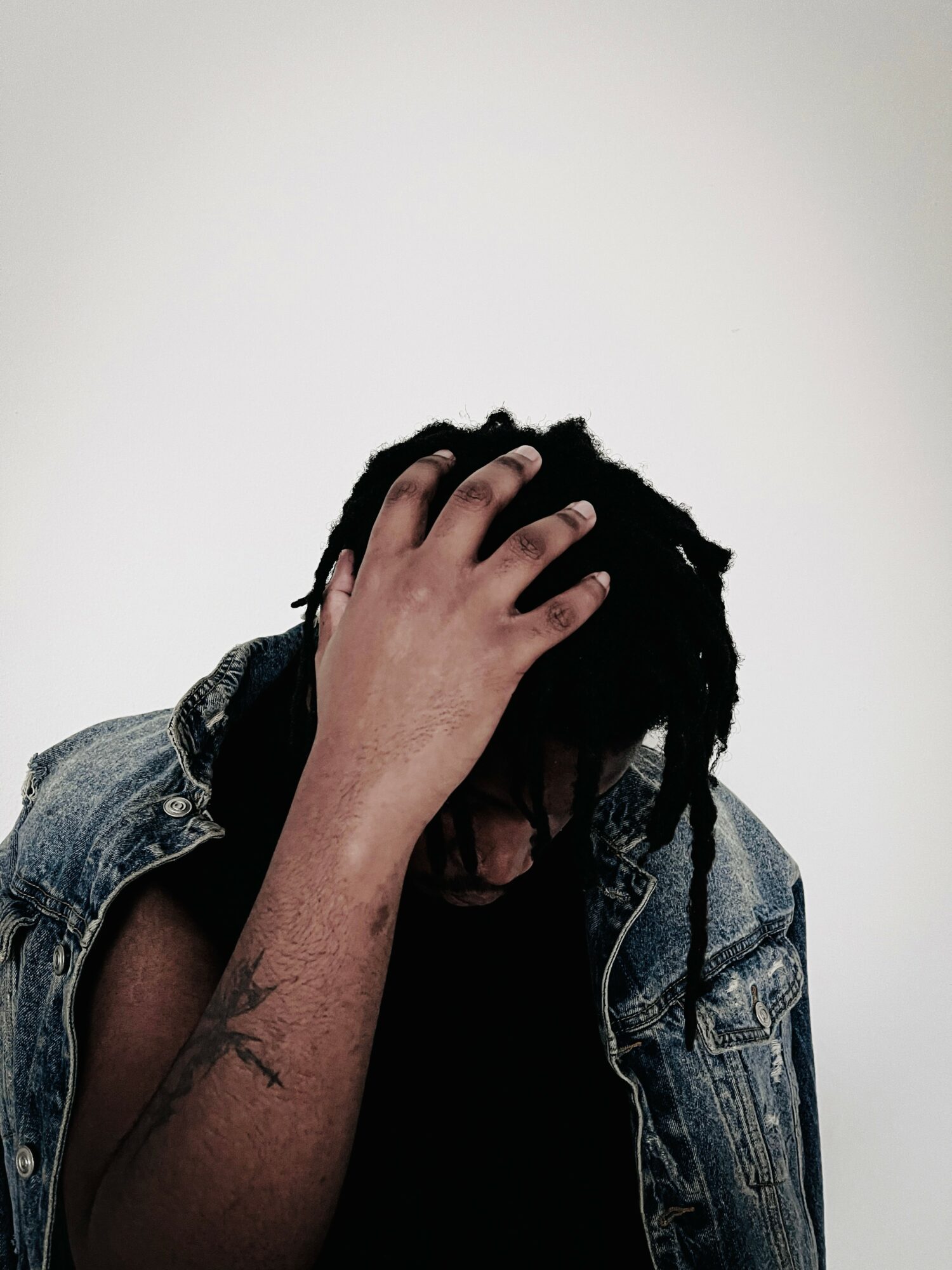 Person with dreadlocks wearing a denim jacket, covering face with hand, against plain background.