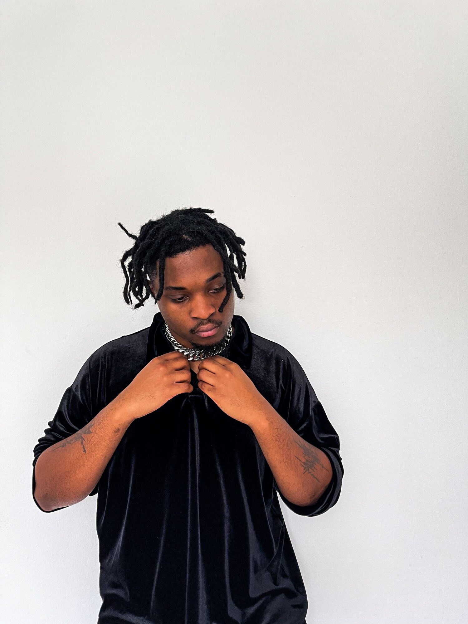 Young man with dreadlocks adjusting his black jacket against a plain white background.