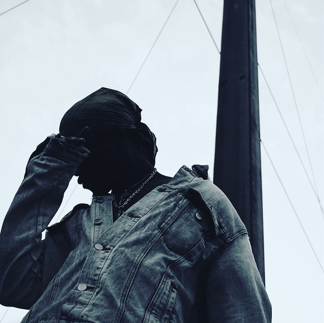 Person with hand on head, wearing denim jacket, silhouette against sky and utility pole, facing left.