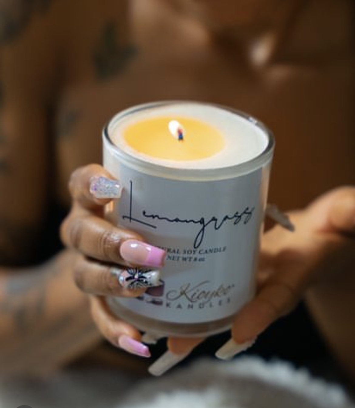 Person holding a lit candle in a glass container with handwritten text, with decorated nails visible.