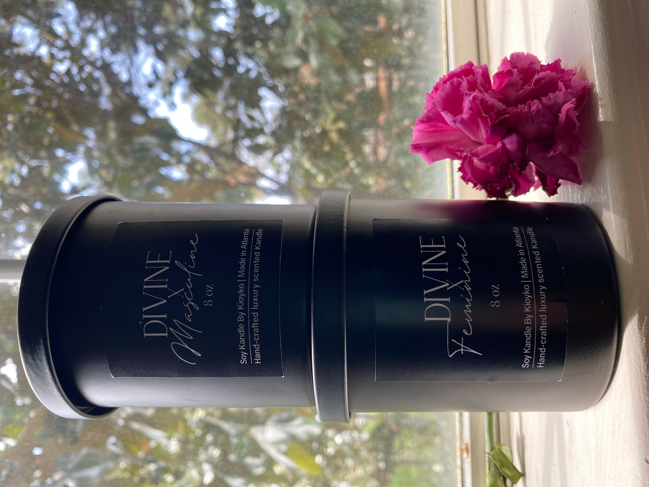 Black cylindrical container with white text, pink flowers on windowsill, trees outside window, sunlight streaming in.