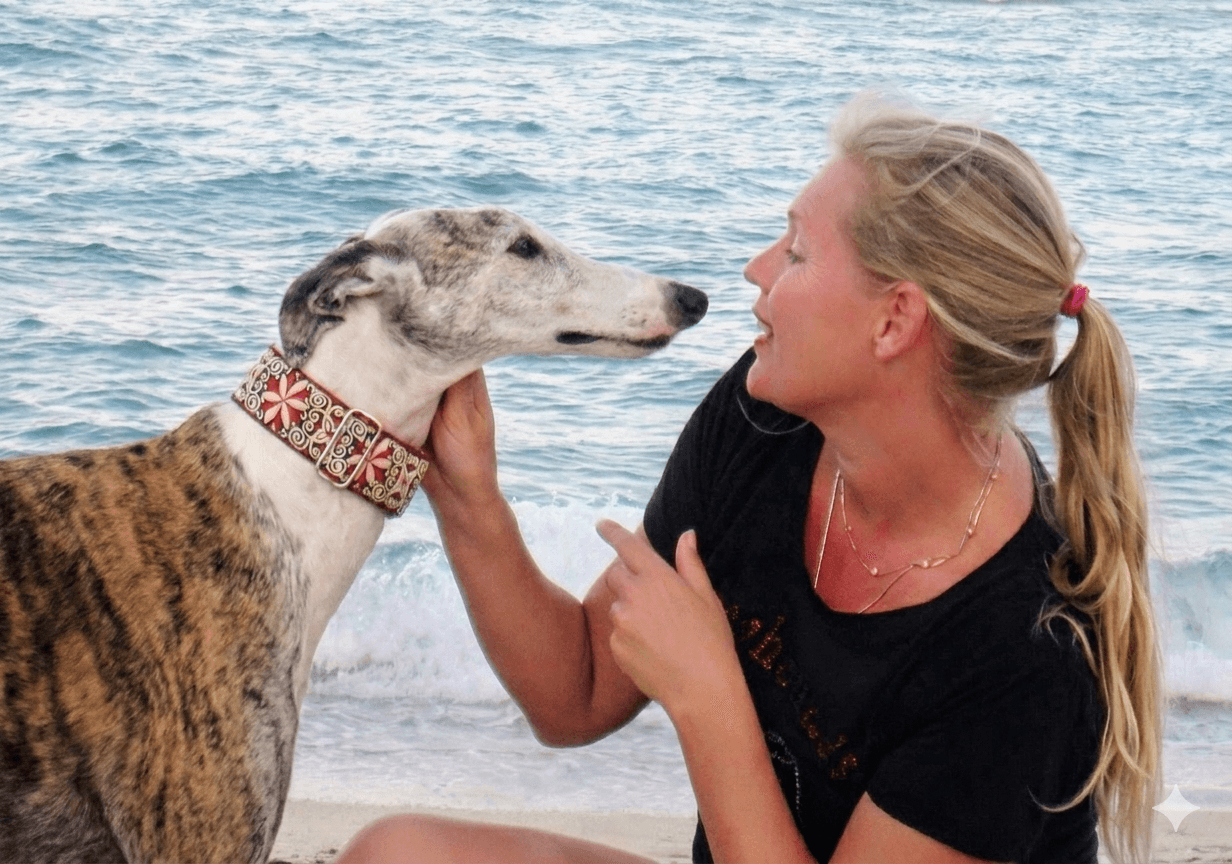 Woman with blonde hair and a black shirt interacts with a large dog on the beach, water in the background.