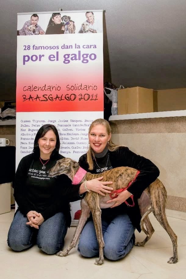 Two women kneel with a dog in front of a poster and a cardboard box, smiling indoors.