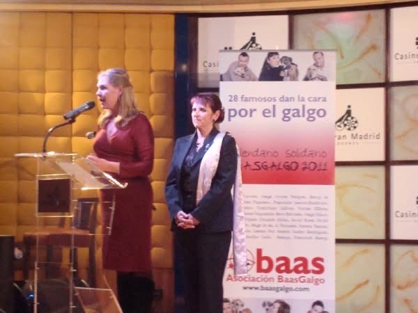 Two women stand on stage, one speaking at a podium with a microphone, the other listening, with a banner behind them.
