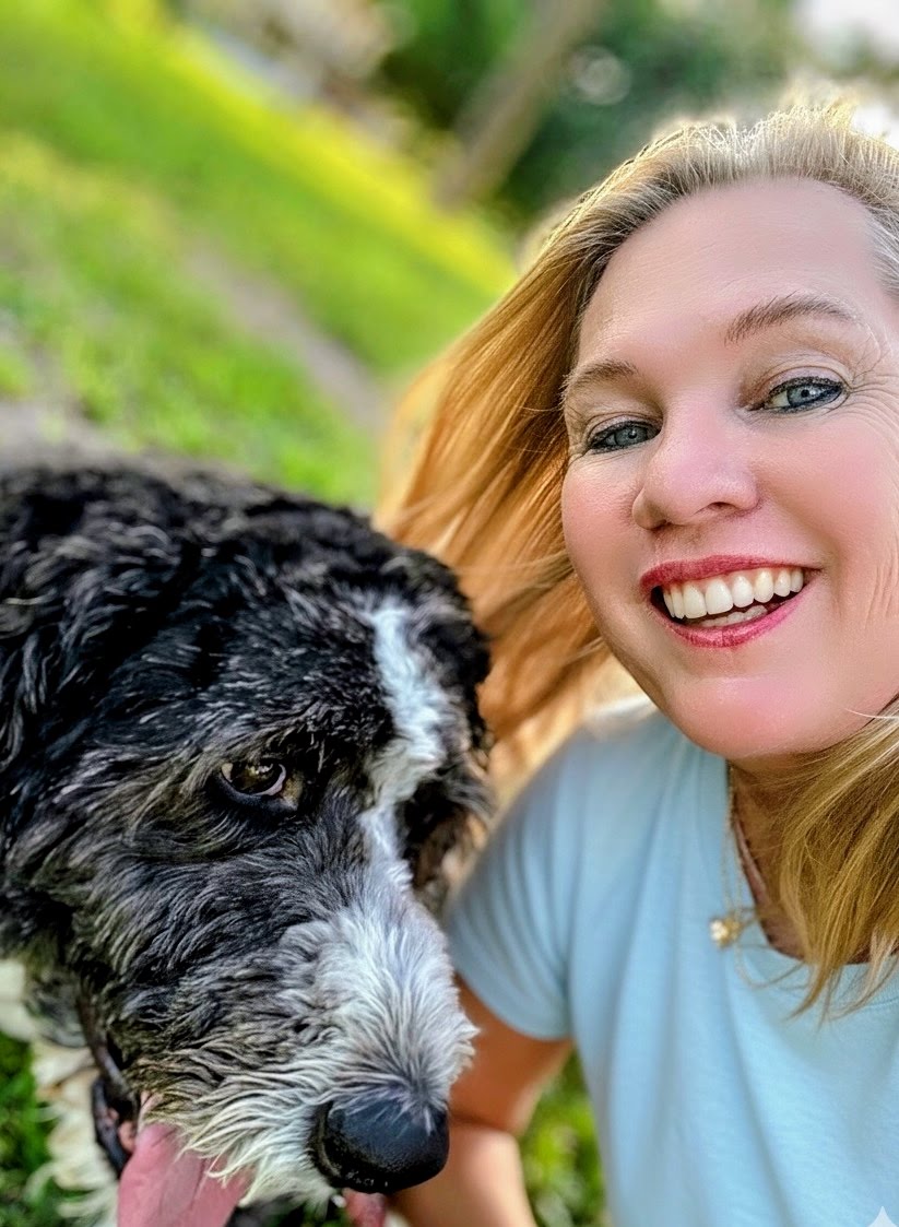 Smiling woman with blonde hair taking a selfie outdoors with a black and white dog, green grass in background.