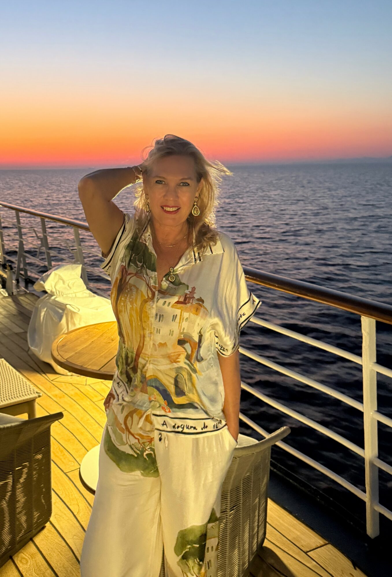 Woman with blonde hair smiling on a ship deck during sunset, ocean in background, wearing a patterned dress.