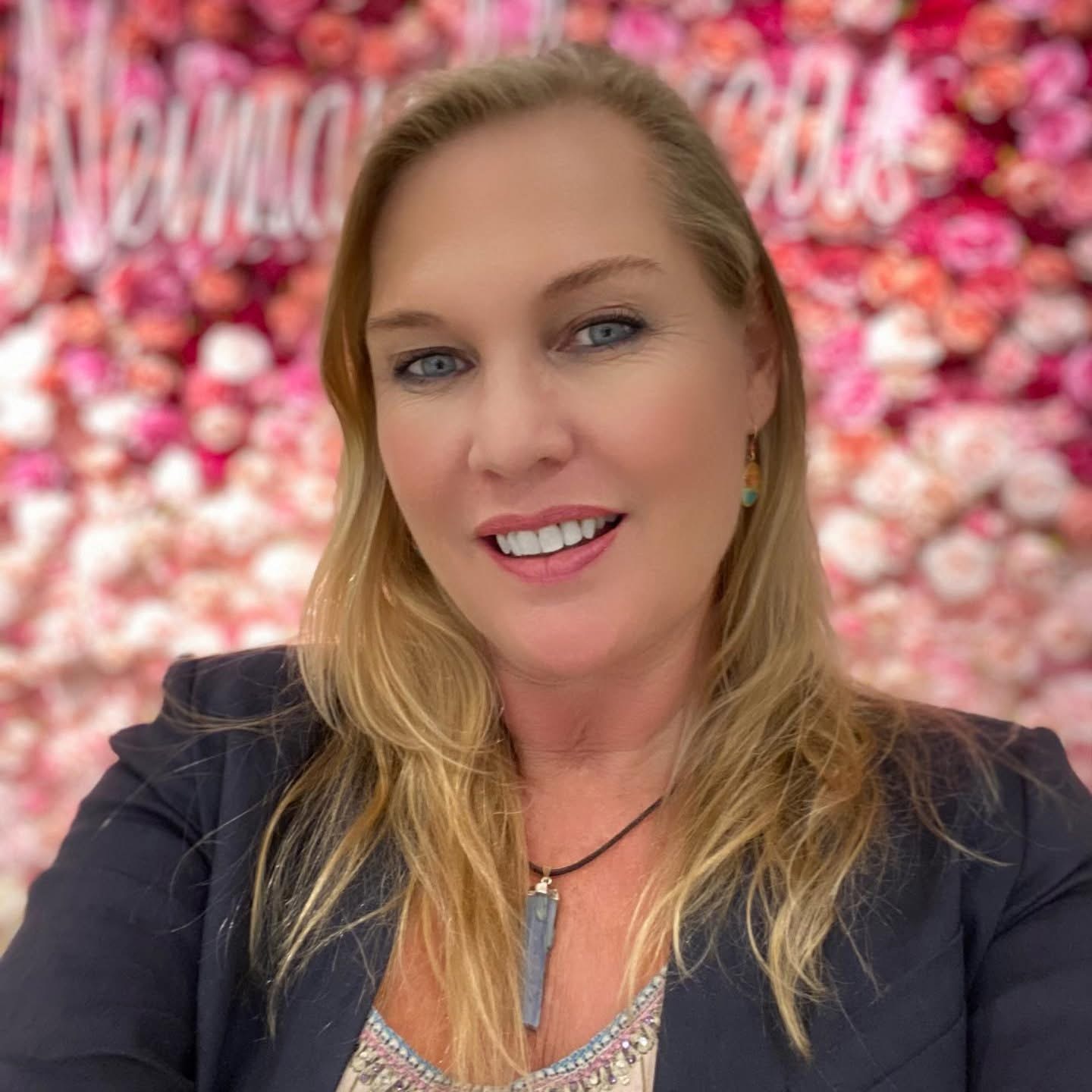 Woman with blonde hair smiling in front of a pink floral background, wearing a dark blazer and a necklace.