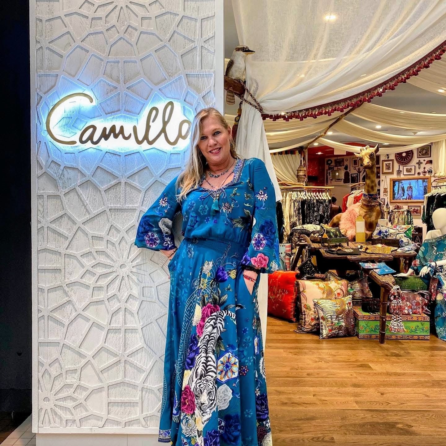 Woman in a blue dress standing in front of a decorative wall with a neon sign that says 'Camilla'.