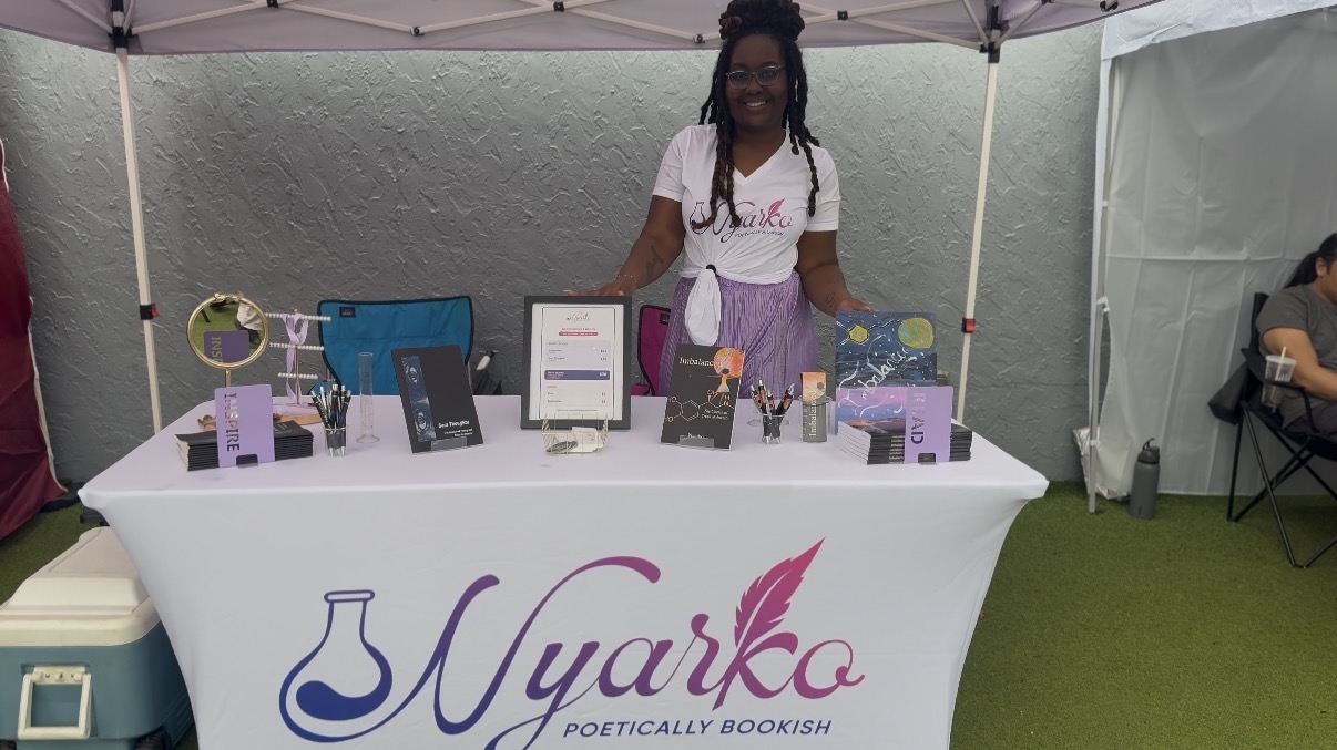 Woman standing behind a table with books and products at a booth, under a canopy, on green grass.