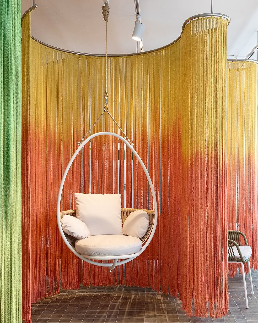 Hanging circular chair with cushions in front of colorful curtain backdrop, with a chair to the right, ceiling and floor visible.