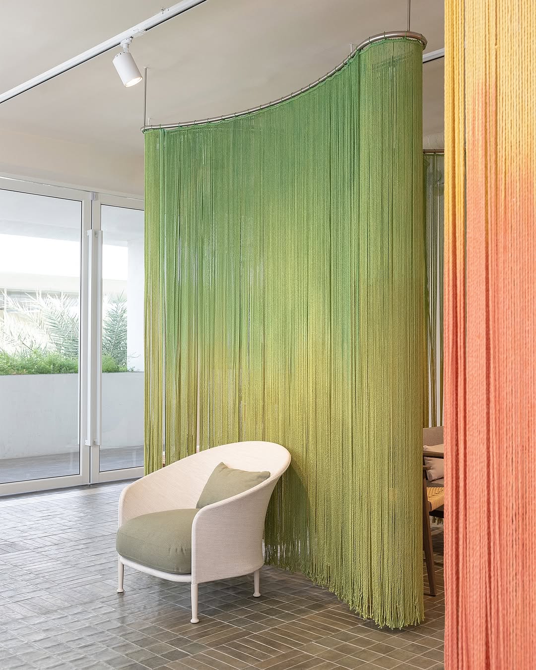 Green beaded curtain separates room from window, with a white armchair nearby, in a bright space with sliding glass doors.