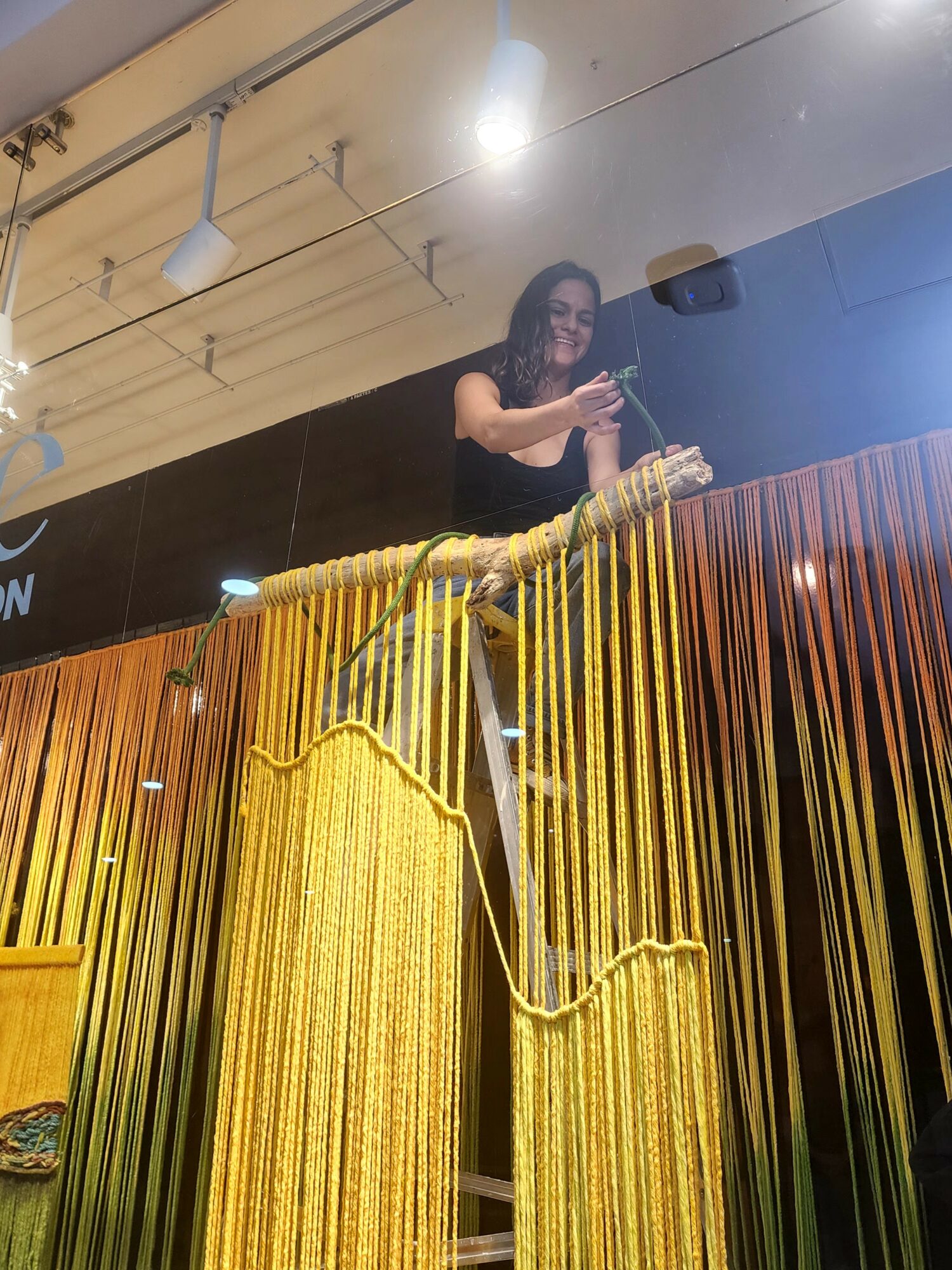 Woman taking a selfie behind a yellow curtain with hanging strands, holding a phone, smiling, in an indoor setting.