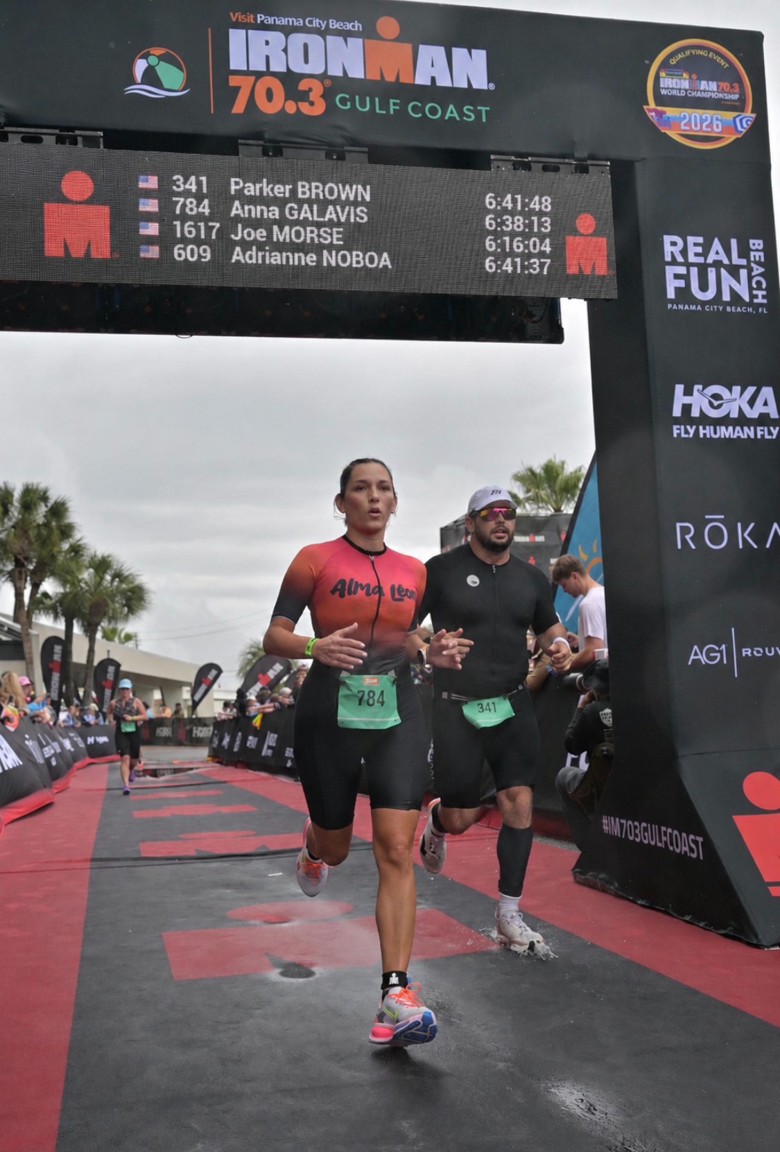 Two runners crossing the finish line at an Ironman event, with a digital timer and event signage overhead.