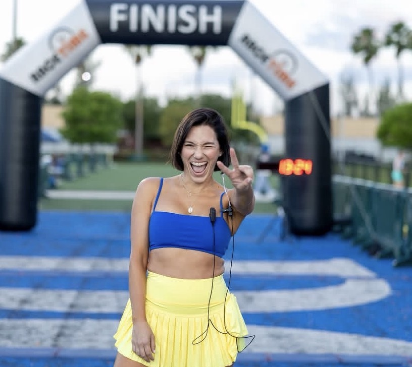 Smiling woman in blue sports bra and yellow skirt at finish line, making peace sign, outdoor setting, trees, blue track.