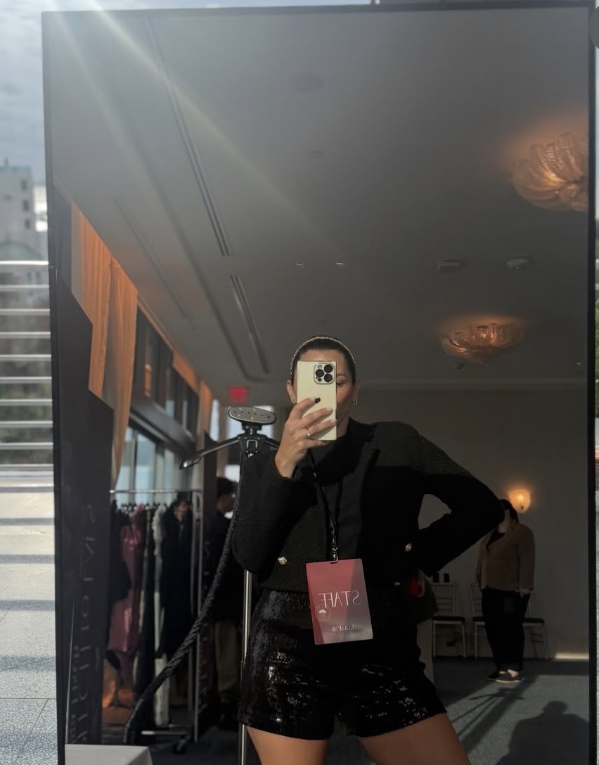 Person taking a mirror selfie indoors, wearing a black outfit with a pink badge, with other people and decor in the background.