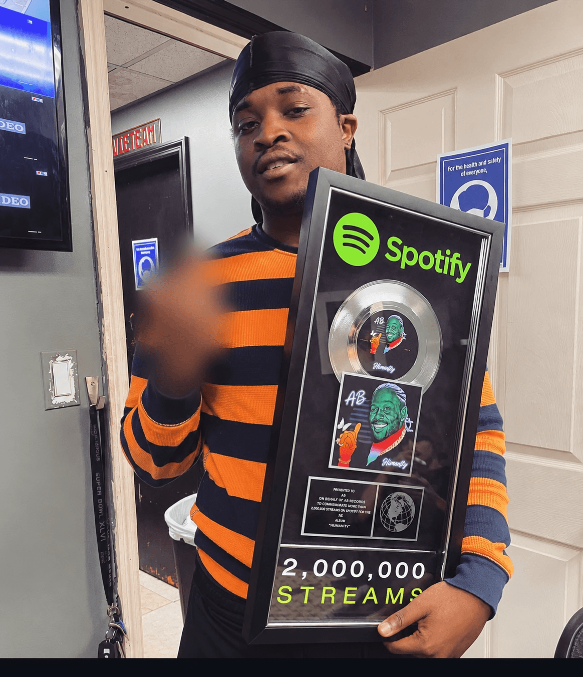 Person holding a framed Spotify award with 2 million streams, wearing a striped shirt and black head covering.