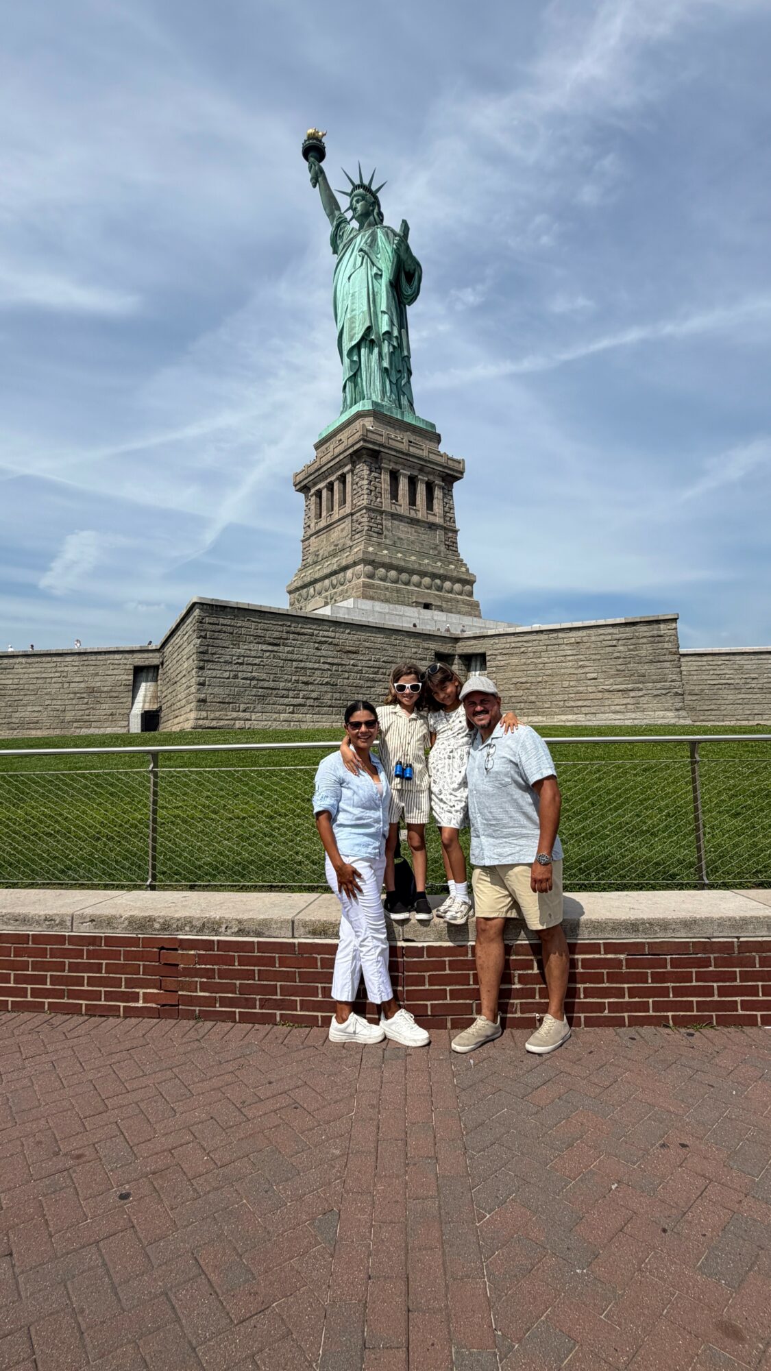 Three people stand in front of the Statue of Liberty on a sunny day, with a green lawn and brick pathway.