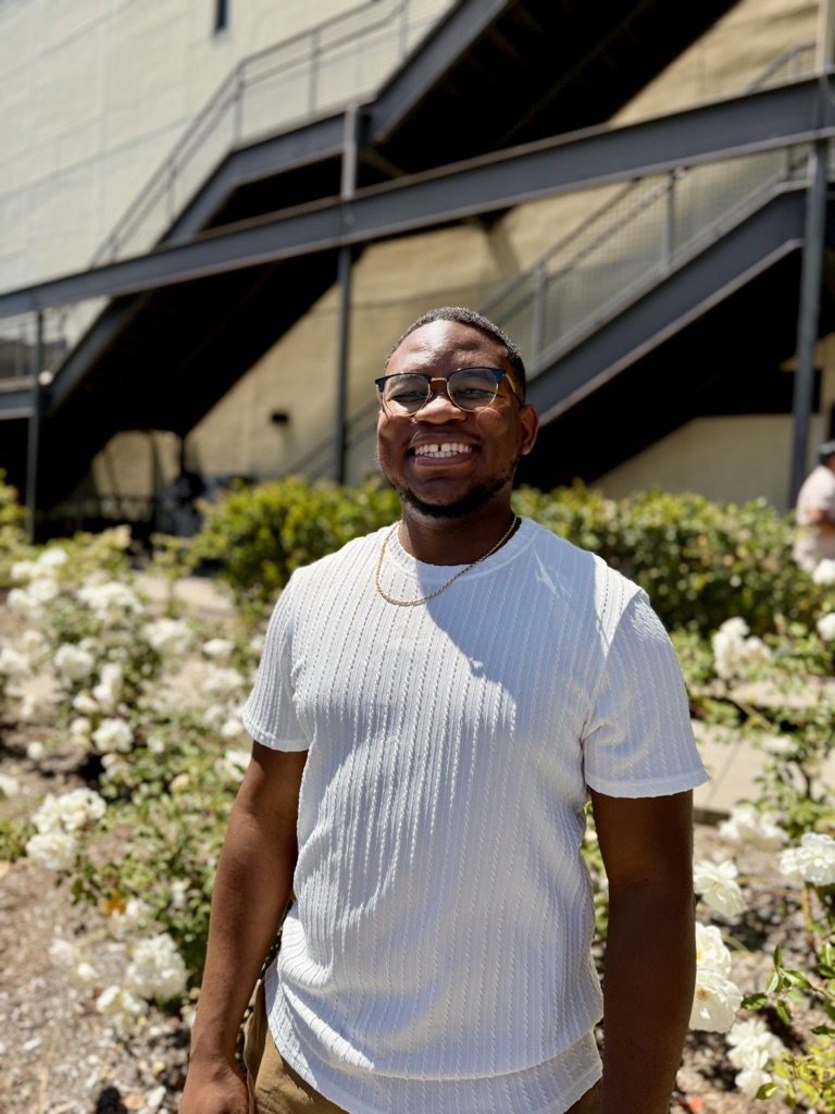 Smiling person wearing glasses and a white shirt outdoors with plants and a building in the background.