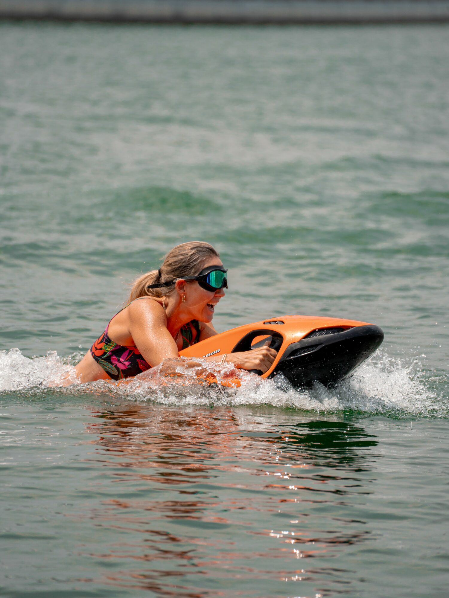 Child wearing goggles rides a watercraft on a body of water, smiling and holding onto the vehicle.
